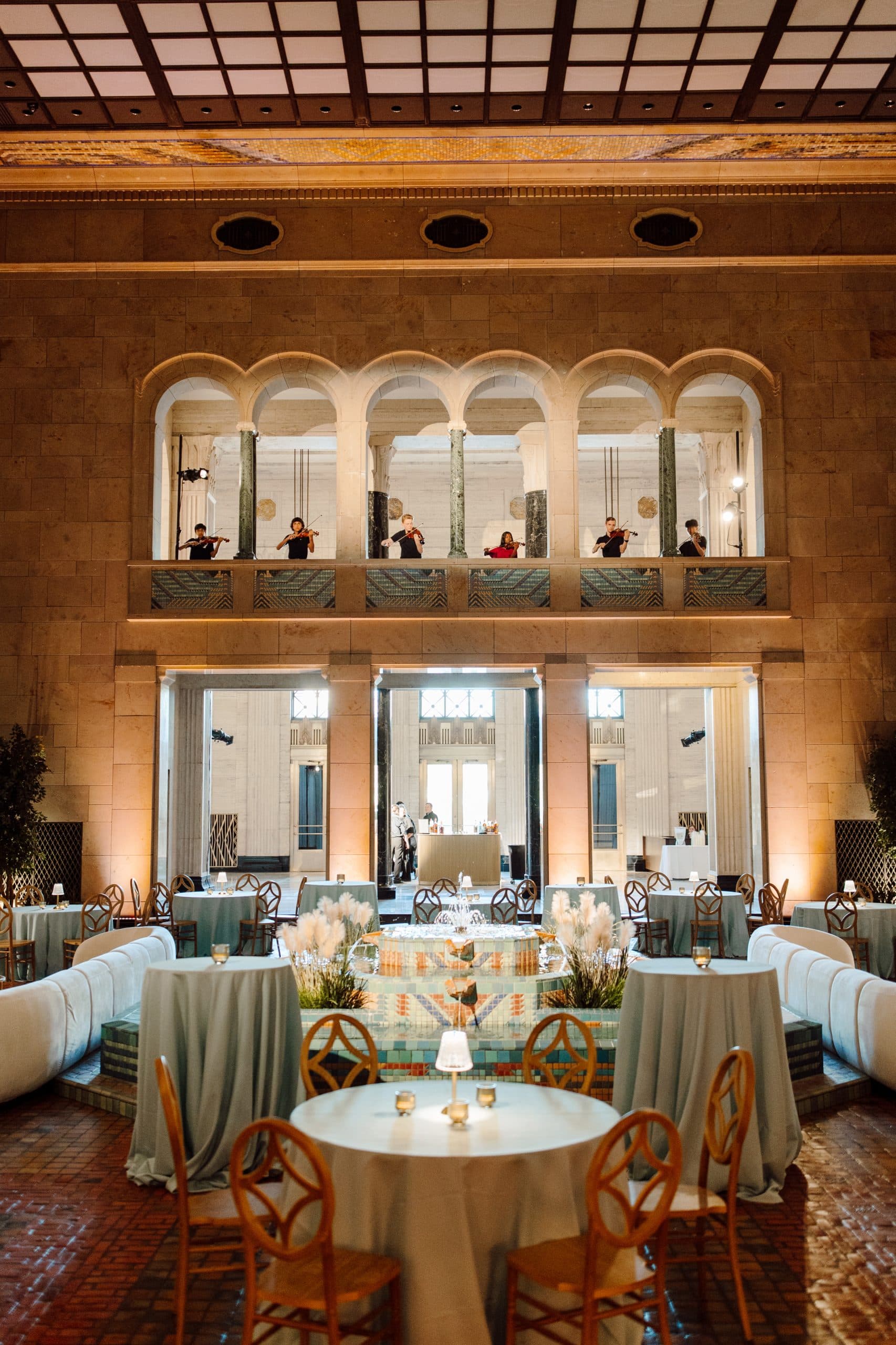 An elegant banquet hall with tall ceilings and an ornate, arched balcony featuring musicians. Round tables with blue tablecloths and wooden chairs are arranged below. Candlelit decorations and soft lighting create a warm ambiance.