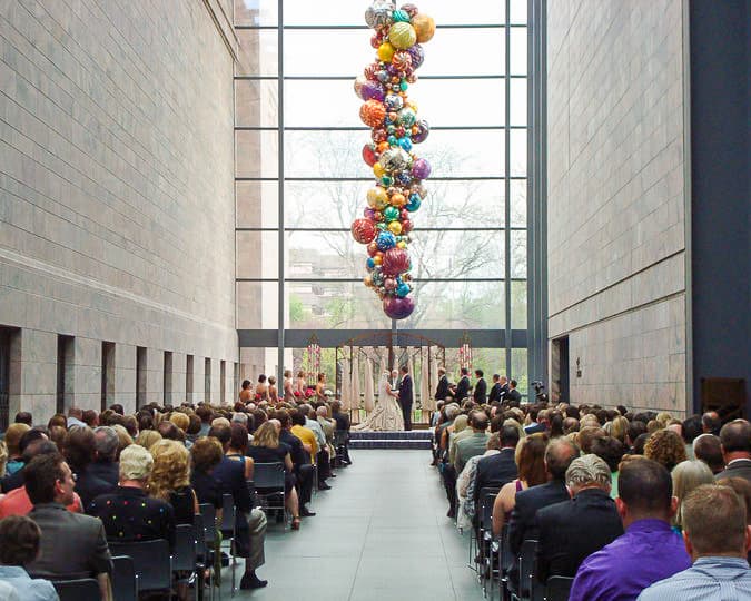 A wedding ceremony takes place in a large hall with a tall glass window. Guests are seated on both sides, facing the couple and officiant. Colorful balloons are suspended from the ceiling, adding a festive touch to the neutral-toned space.