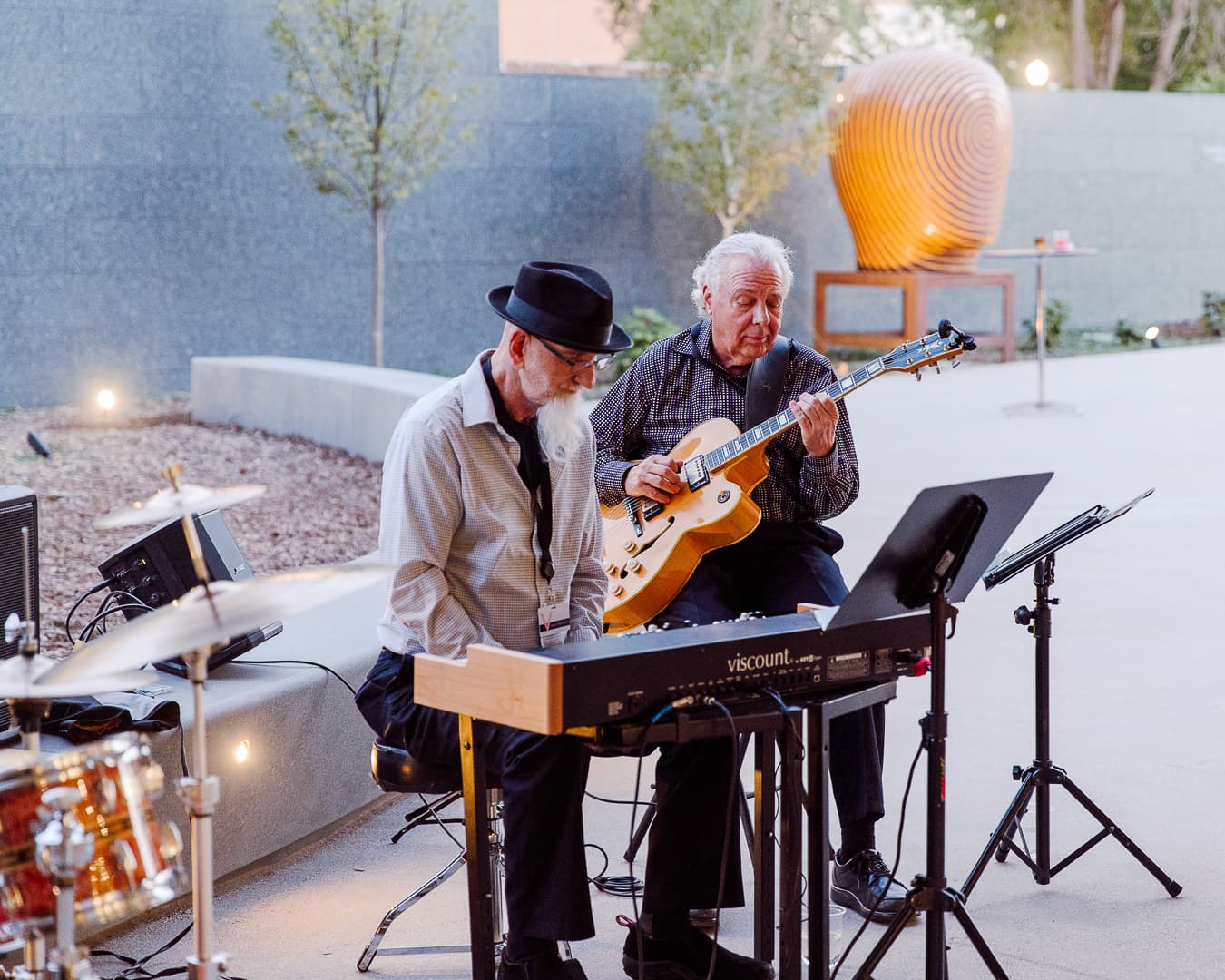 Two elderly men are performing music outdoors. One is playing a keyboard and wearing a hat, and the other is playing a guitar. They are seated near music stands, with trees and a sculpture in the background.