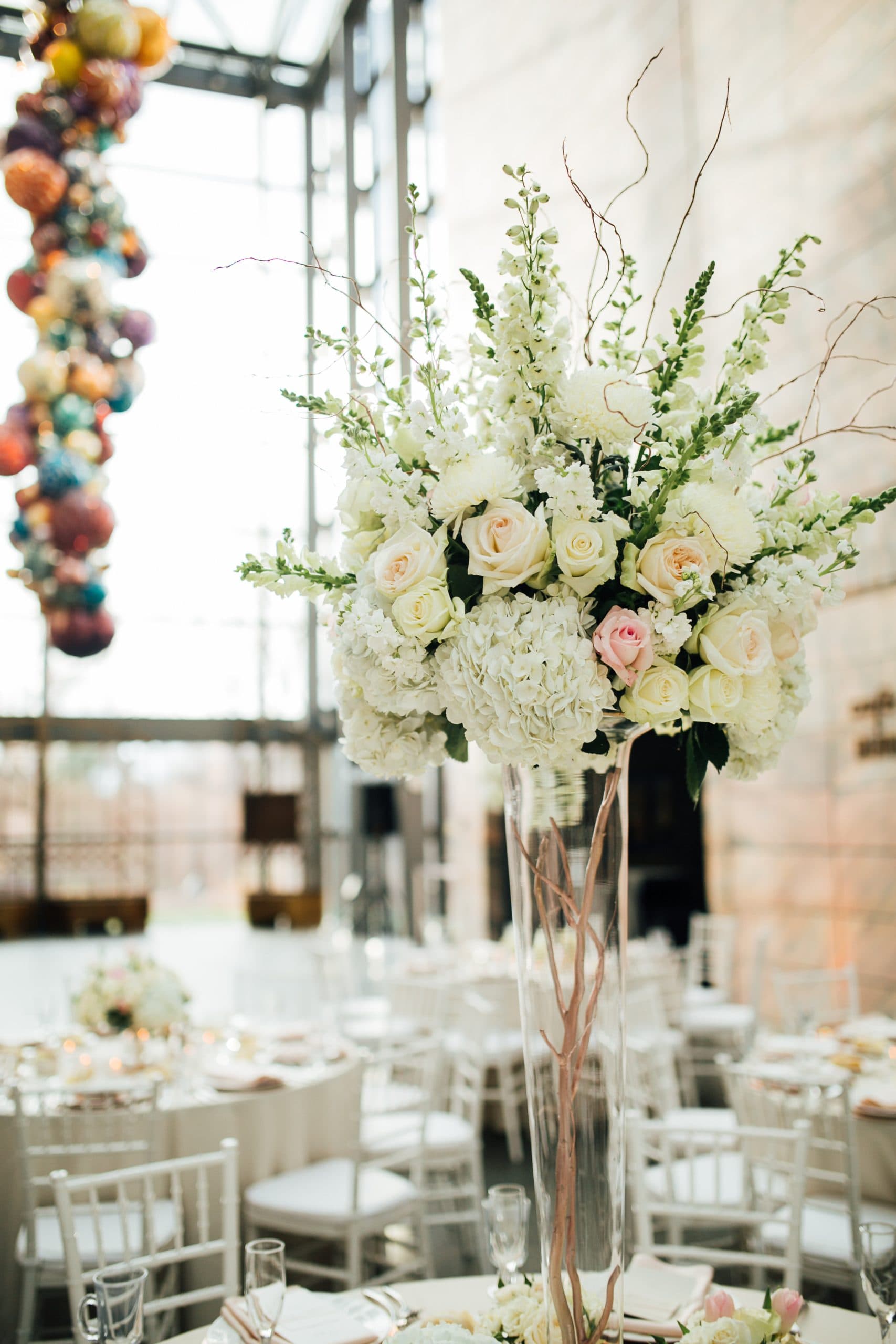 Tall floral centerpiece with white roses and hydrangeas, accented by curly branches in a glass vase. Surrounding are elegantly set tables with white chairs. A colorful glass chandelier hangs in the background.
