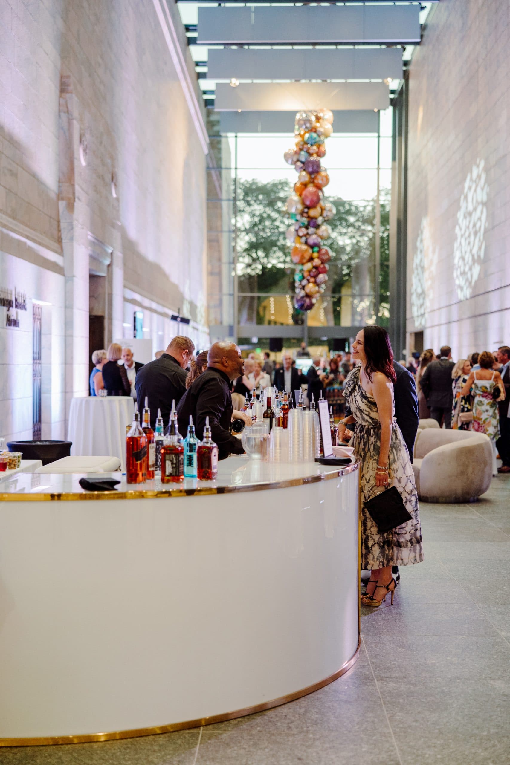 A stylish indoor event with a modern bar in the foreground, stocked with a variety of colorful drink bottles. People are mingling and talking in the background. A striking ceiling installation of suspended colorful spheres is visible above.