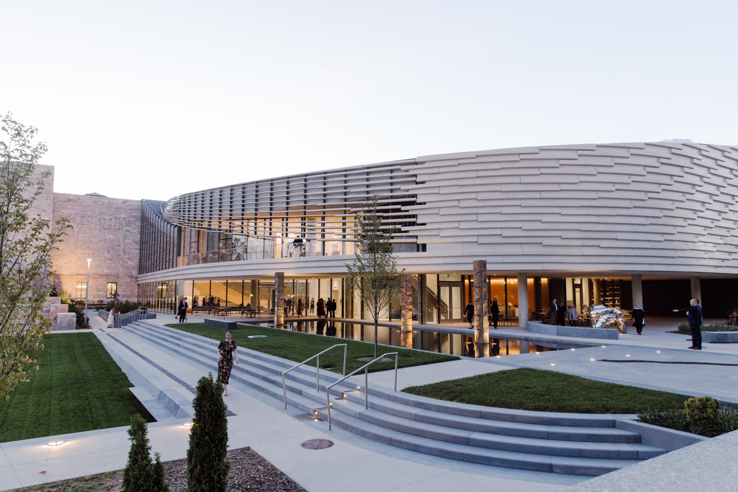 Modern building with a distinctive, wavy design, featuring large glass windows. The entrance is surrounded by landscaped gardens and steps. People are gathered near the entrance, and the scene is set during early evening with a clear sky.