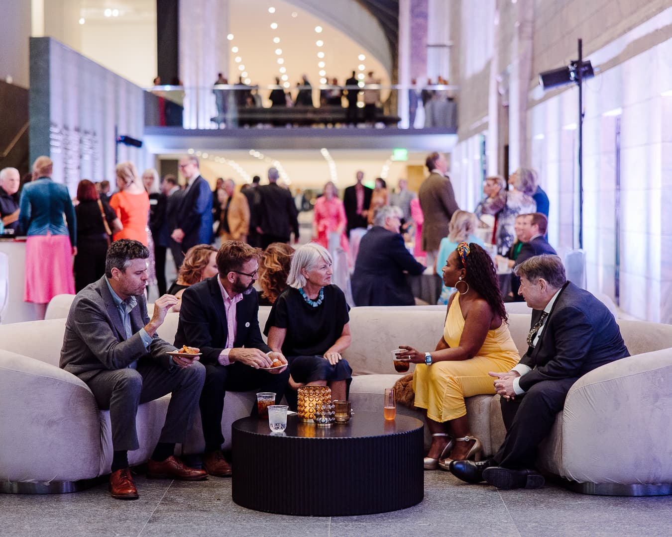 A group of five people sit on a curved sofa at a formal event, engaged in conversation. They are surrounded by other guests mingling in a spacious, elegantly lit venue. Decorative candles and drinks are on the small table in front of them.