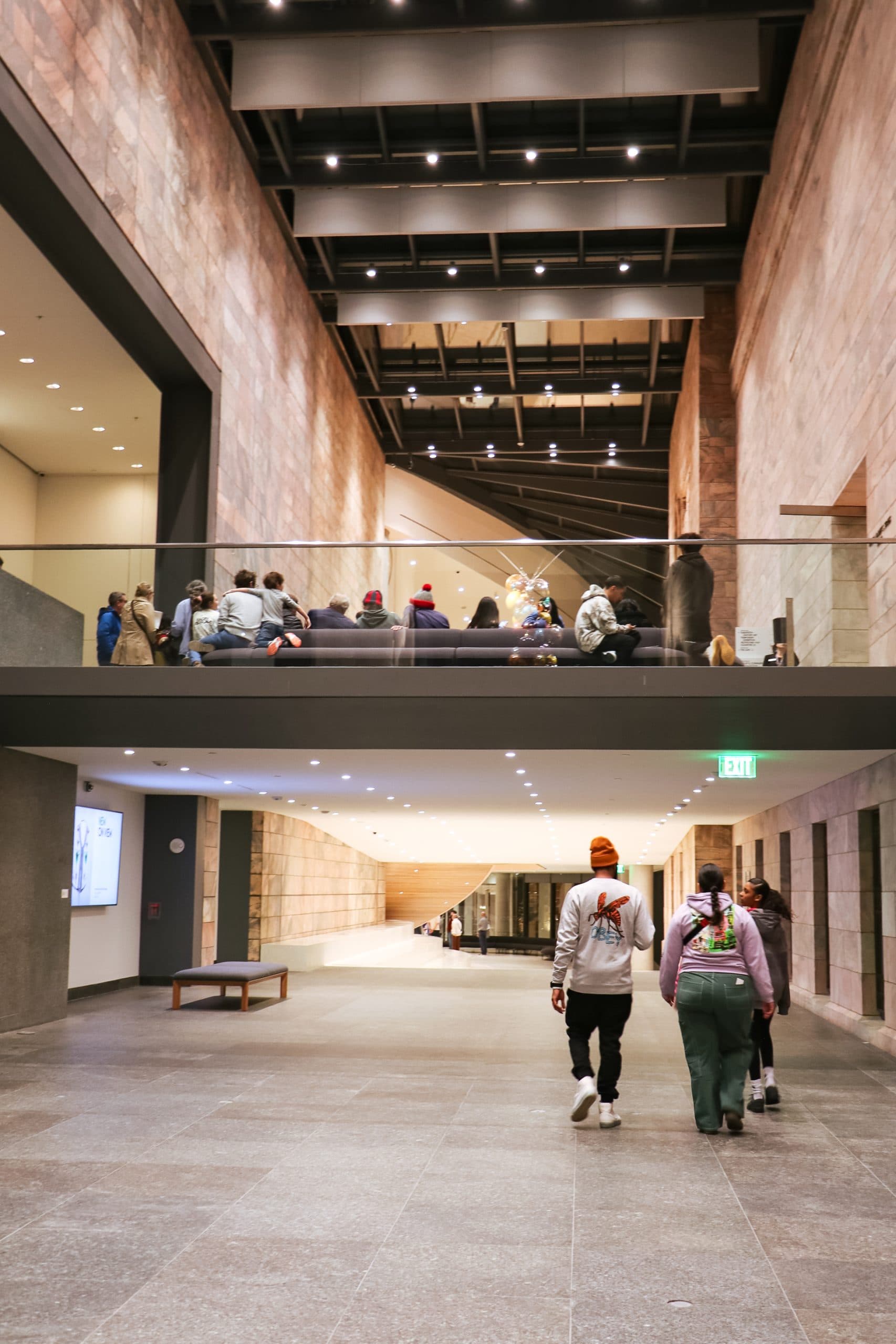 People walk through a modern, spacious hallway with high ceilings and stone walls. Others are seated on a bench on an upper level. A display with lighting is visible in the background. An exit sign glows green.