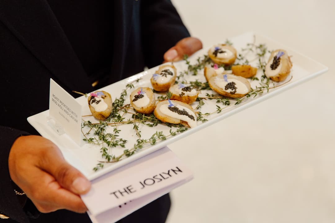 A person holds a rectangular tray with small appetizers topped with cream and garnishes, including caviar and herbs. The tray rests on a bed of thyme. A card and a paper labeled "THE JOSLYN" are visible beneath the tray.
