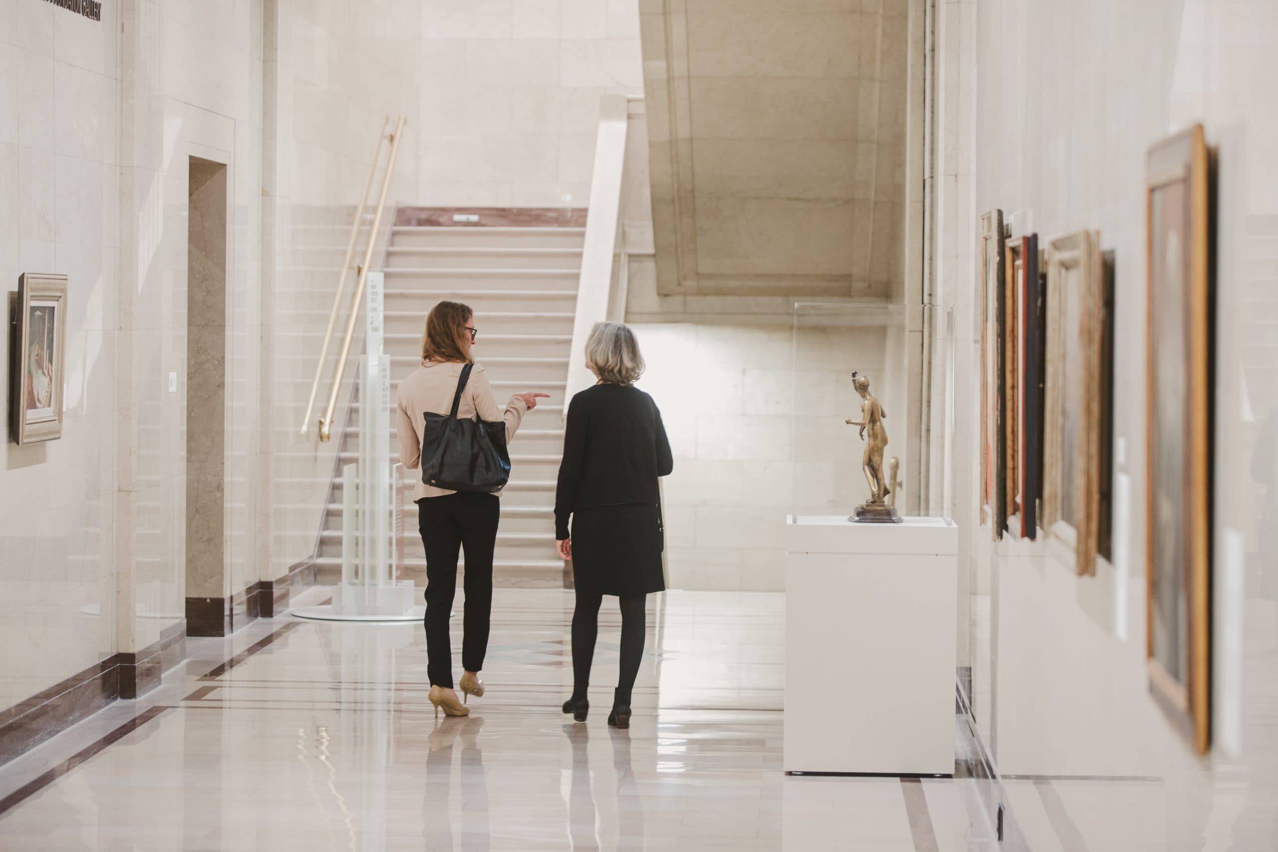 Two women walk down a bright, marble hallway in an art museum, engaged in conversation. Paintings hang on the walls and a small sculpture is displayed on a pedestal to their right.