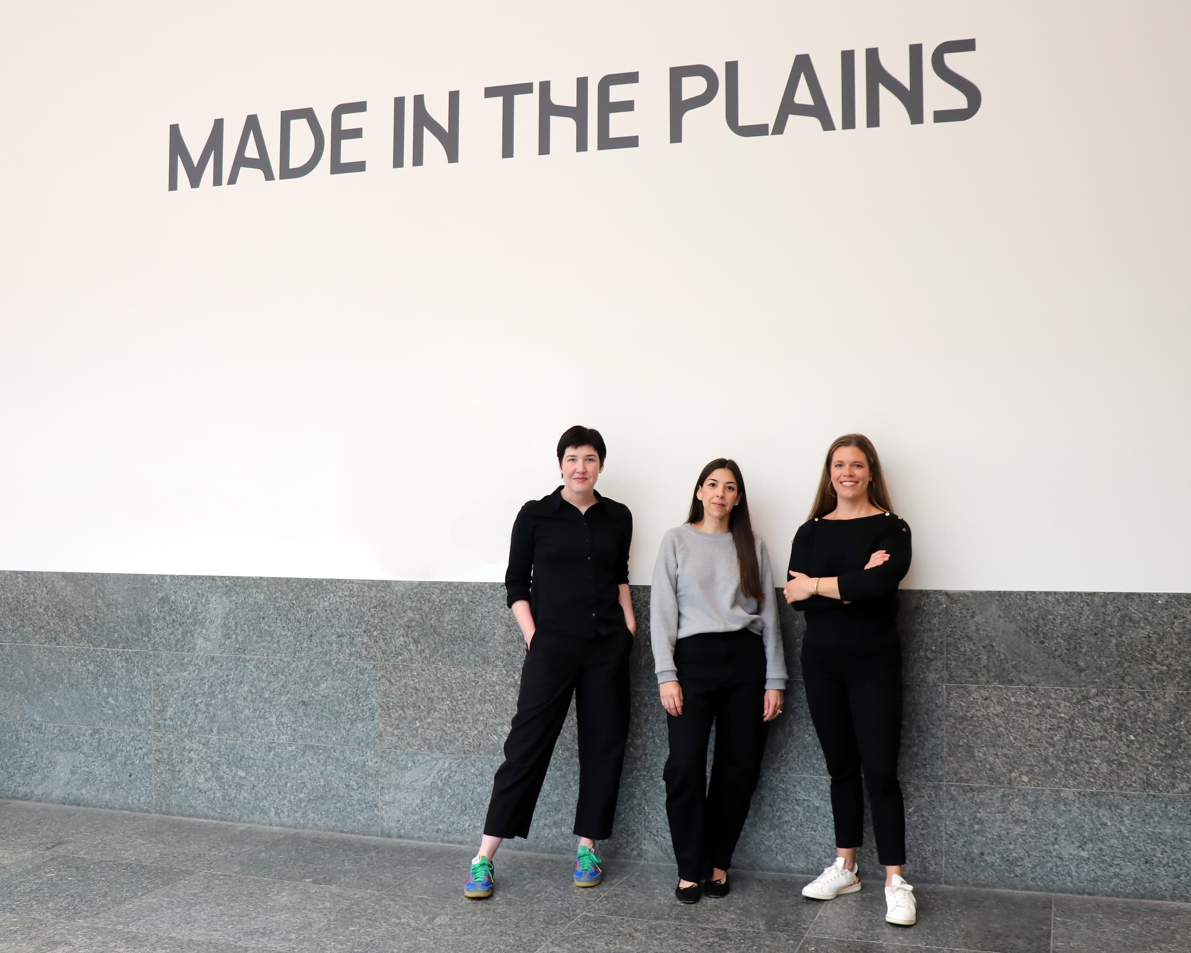 Three women stand side by side, smiling, in front of a wall with the large text "MADE IN THE PLAINS." The setting appears to be indoors, with a grey tiled floor and wall.