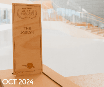 A wooden award plaque titled "THE JOSLYN" with "LAURELS AWARDS 2024" engraved, placed on a table. The background shows a modern staircase. "OCT 2024" is written in white in the bottom corner.