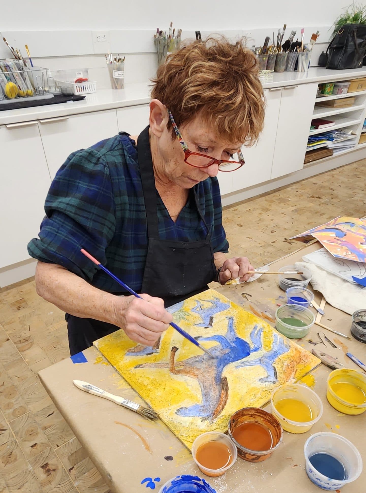 An older woman wearing glasses and a dark apron paints a colorful abstract design on a canvas at a table, surrounded by jars of paint in shades of blue, brown, yellow, and green in an art studio.