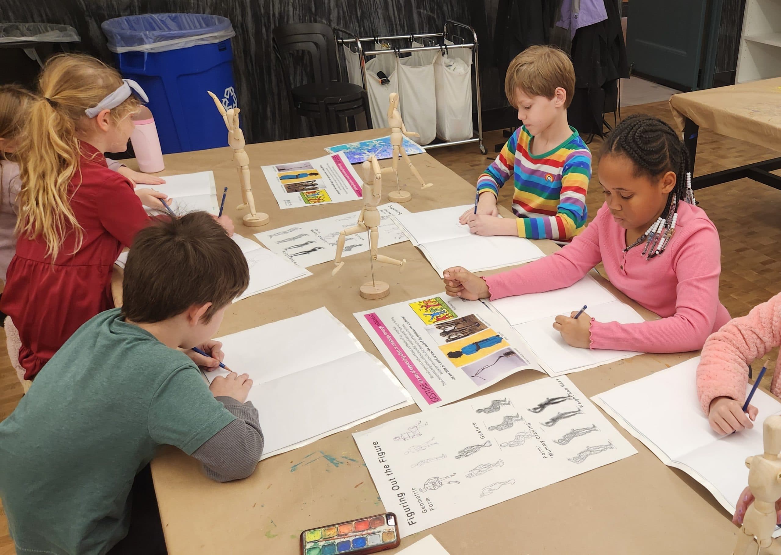 Five children sit around a table drawing on white paper with pencils. Wooden artist mannequins, drawing guides, and watercolor sets are on the table, indicating an art class setting.