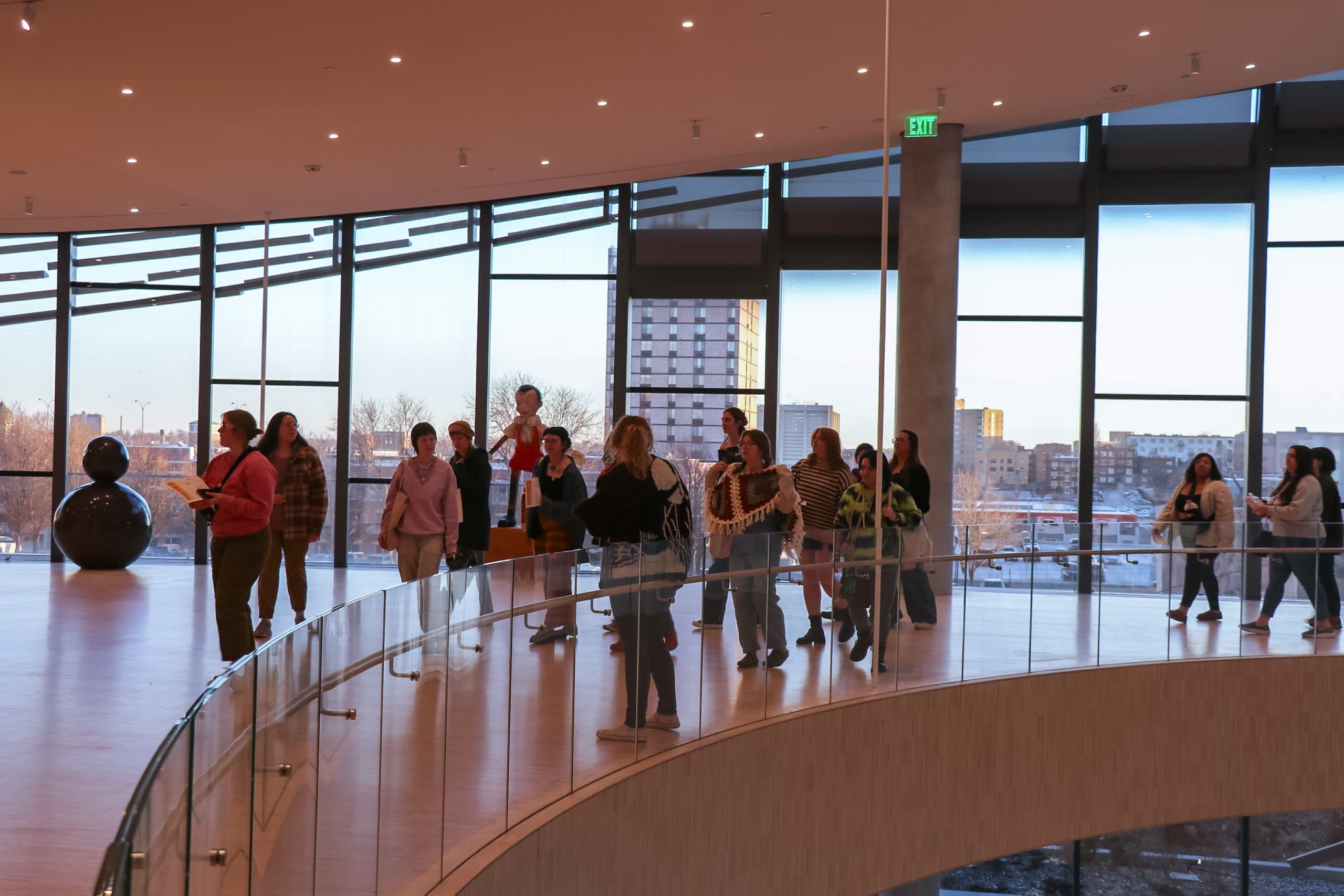 A group of people stand and walk along a curved indoor balcony with large glass windows overlooking a cityscape at sunset. Some are talking or taking photos. The area is spacious and modern.
