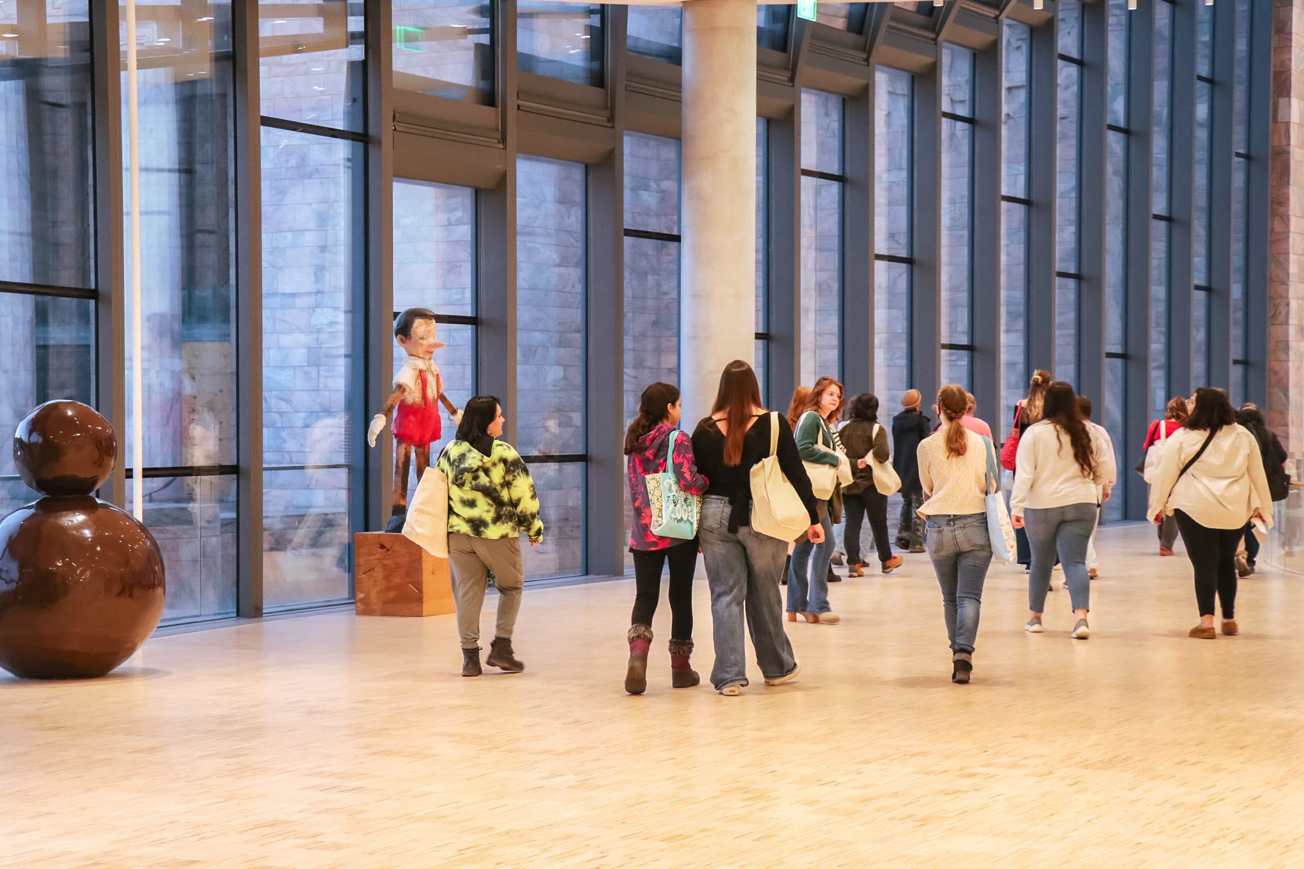 A group of people walk through a spacious, modern gallery with large windows and sculptures, including a brown abstract figure and a colorful statue near the glass wall. Most people carry bags and are looking around.