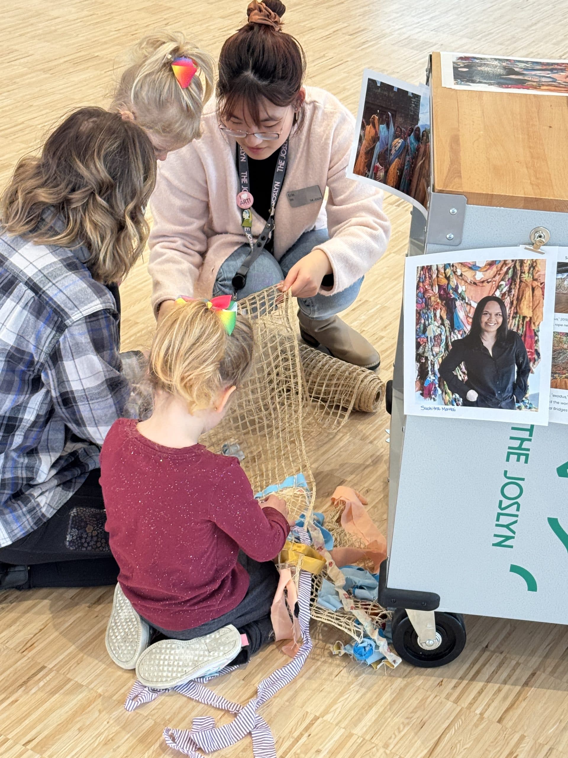 Three children and an adult sit on a wooden floor, weaving or crafting with netting and colorful fabric strips. A cart with photos and the word "Joslyn" is nearby. They appear to be engaged in an art activity.