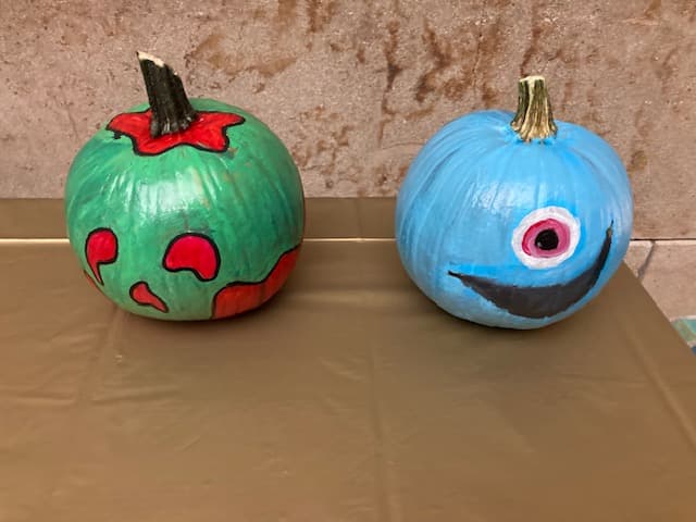 Two painted pumpkins sit on a brown table. The left pumpkin is green with red paint resembling a spooky face. The right pumpkin is blue with a large single eye and a smiling mouth, creating a monster-like appearance.