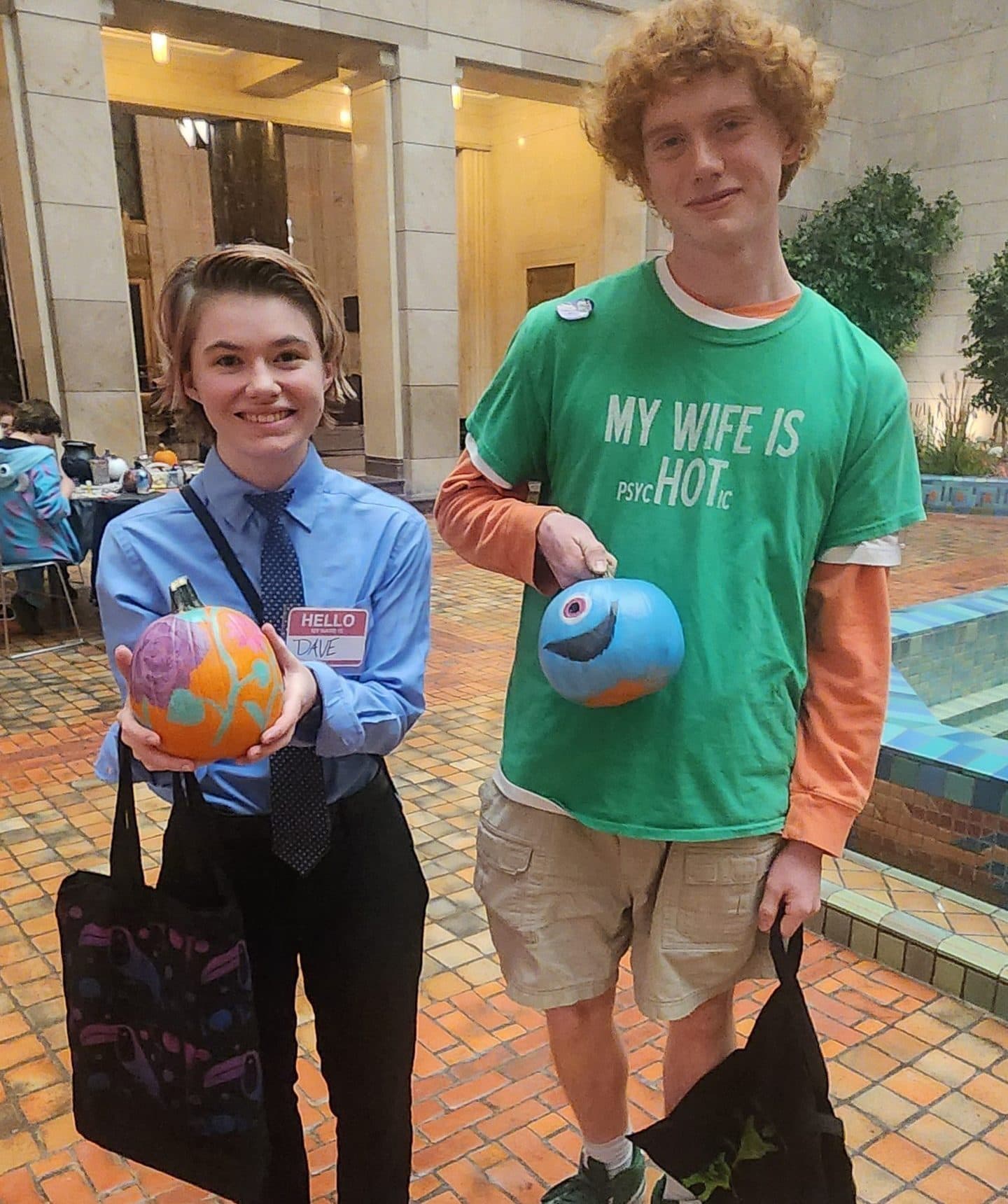 Two people stand indoors on a brick floor, smiling and holding colorful painted ceramic objects. One wears a blue shirt, tie, and name tag; the other wears a green T-shirt, shorts, and holds a black tote bag.