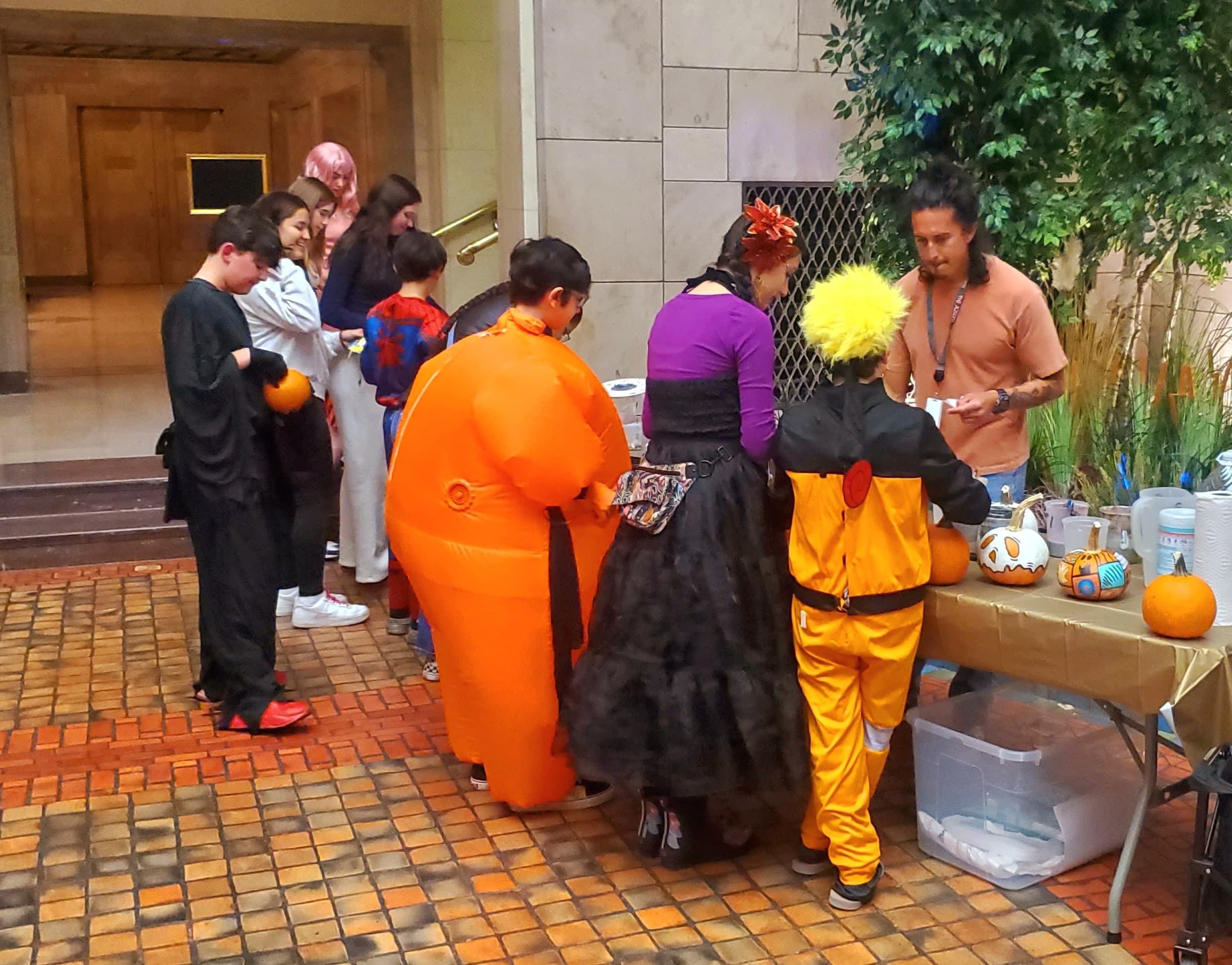 A group of people in colorful costumes stand in line at a table indoors, participating in a Halloween or cosplay event. Decorations and supplies are visible on the table, and pumpkins are displayed nearby.