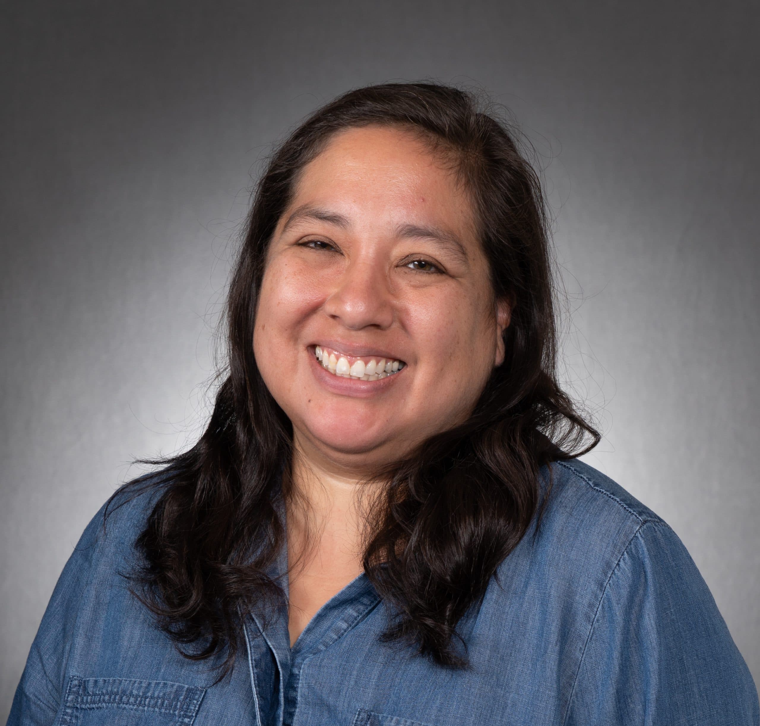 A woman with long dark hair, wearing a blue denim shirt, smiling broadly in front of a plain gray background.