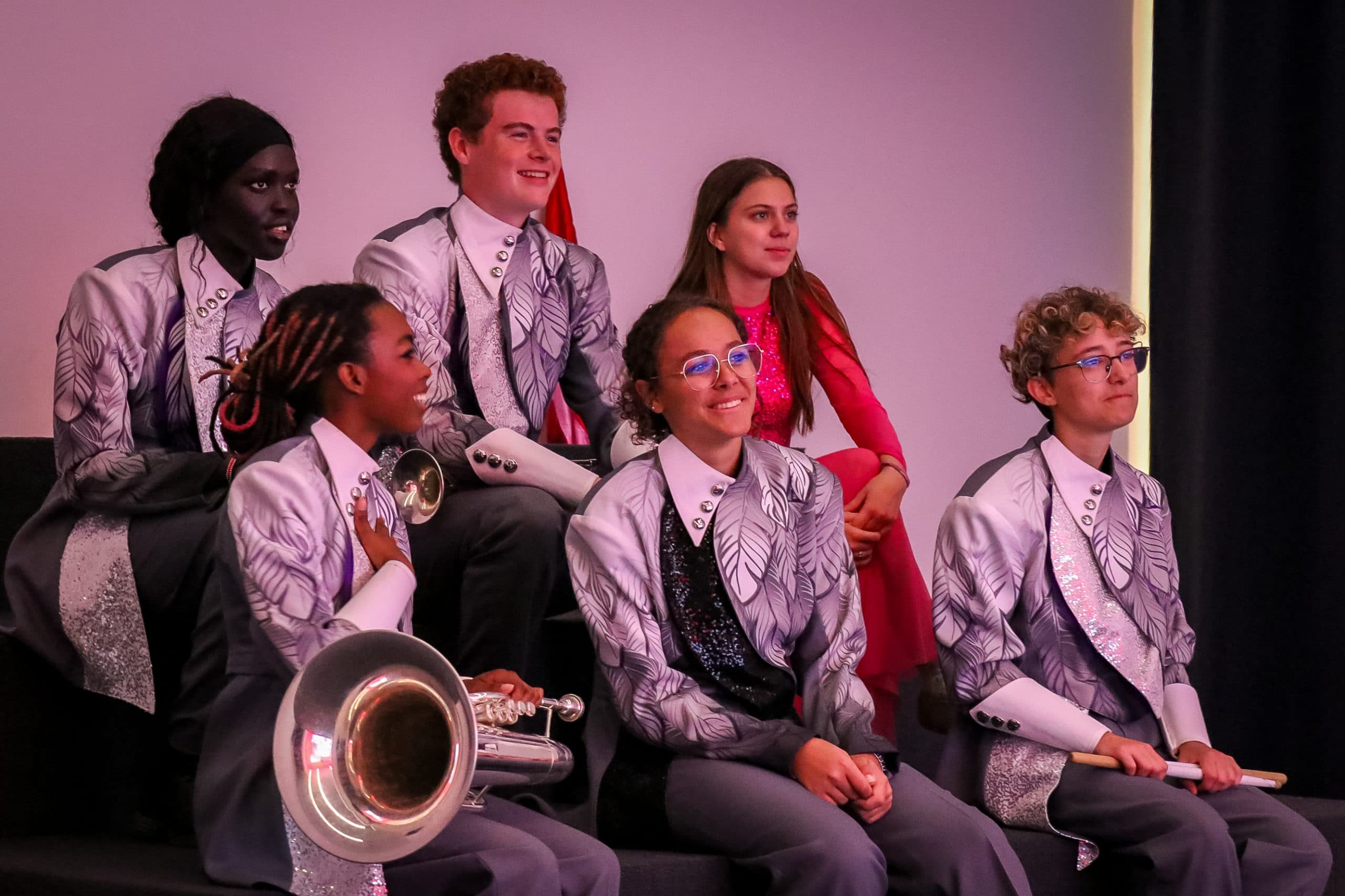 Six young people sit together indoors. Five wear matching silver marching band uniforms and hold instruments, while one, dressed in red, sits behind them. They all appear relaxed and content, posing for a group photo.