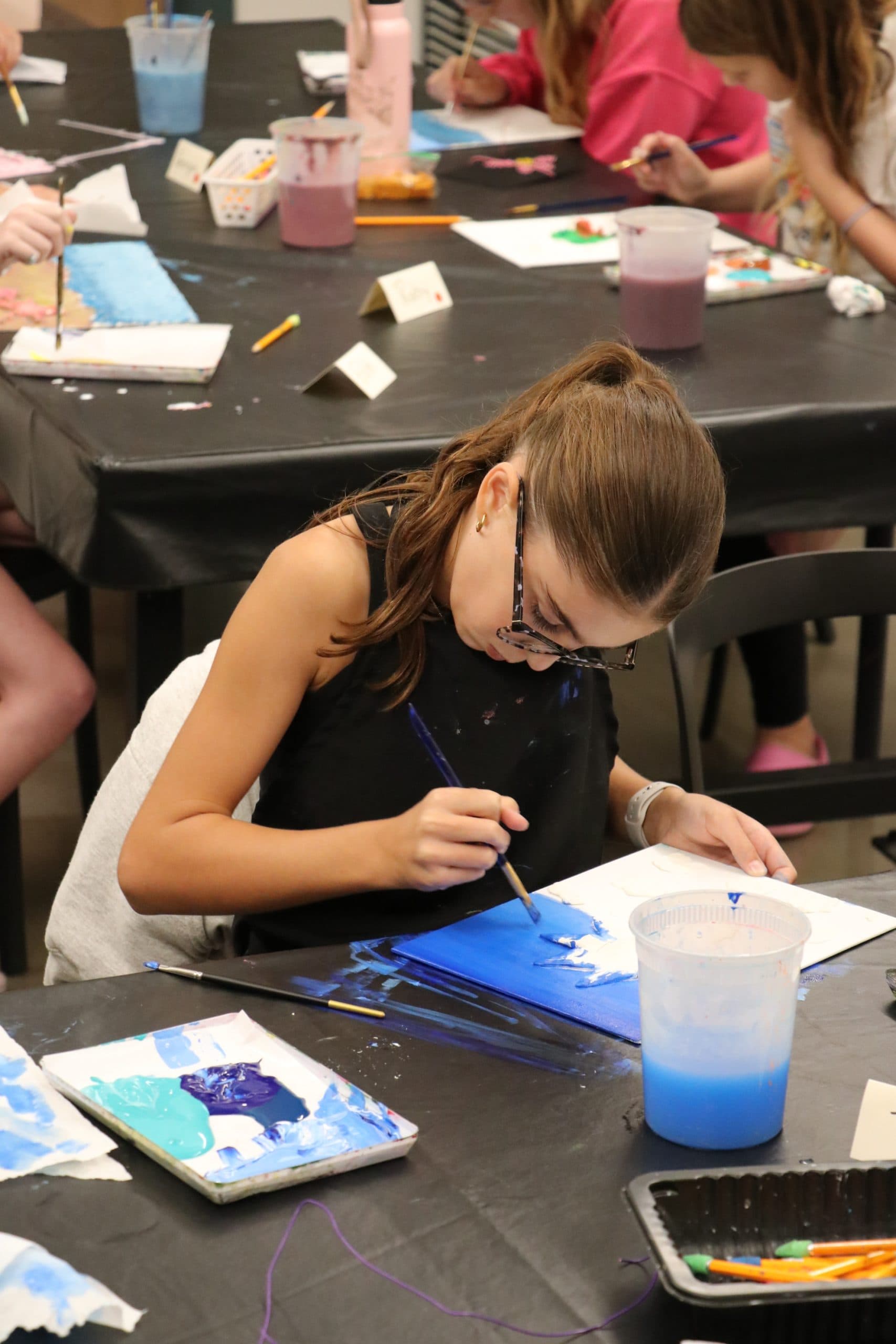 A girl wearing glasses paints on a canvas at a table covered with art supplies, surrounded by other children who are also painting.