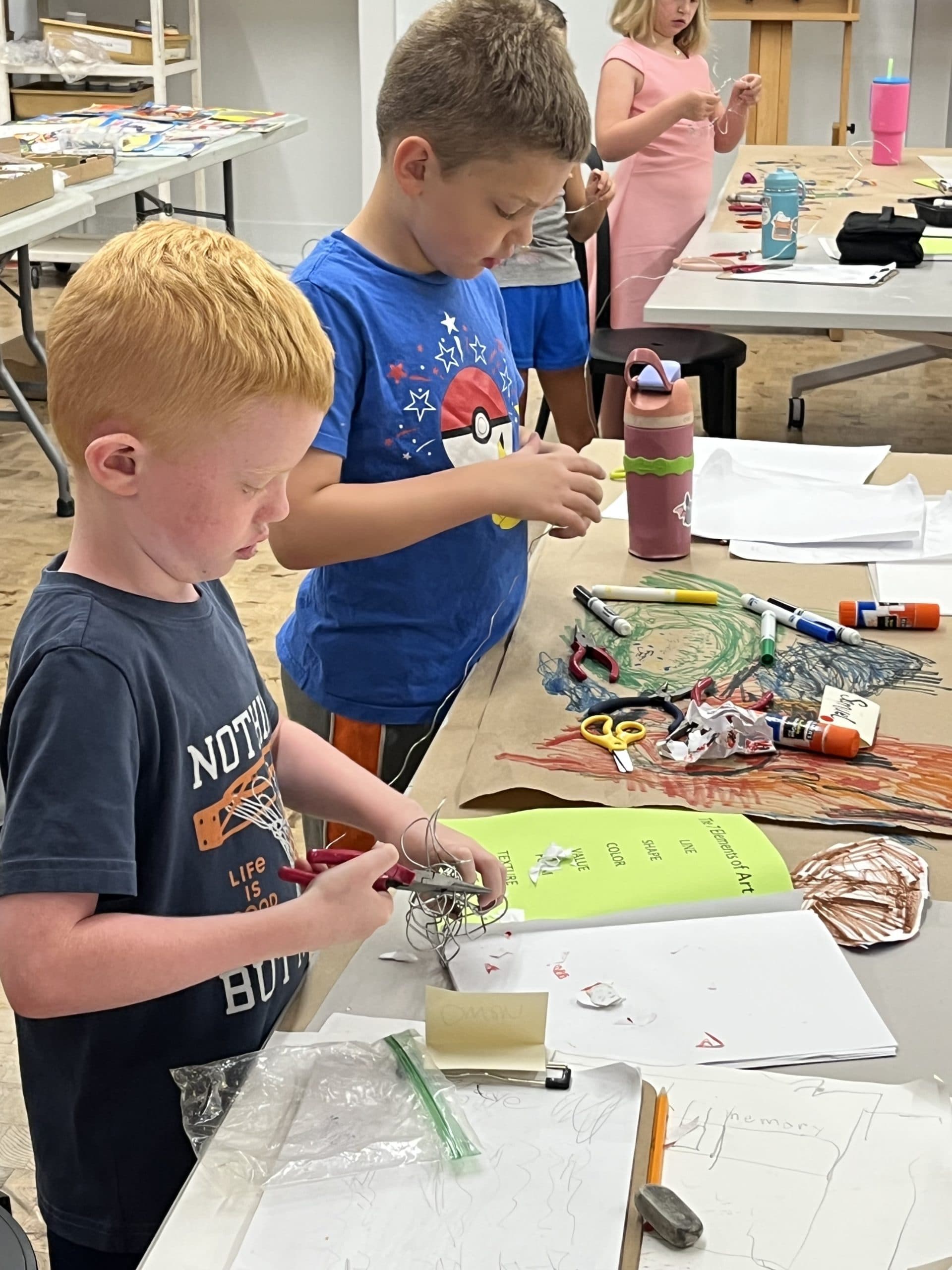 Two young boys stand at a table covered with art supplies, such as glue, scissors, paper, and markers, focused on their creative projects. Other children and finished crafts are visible in the background.
