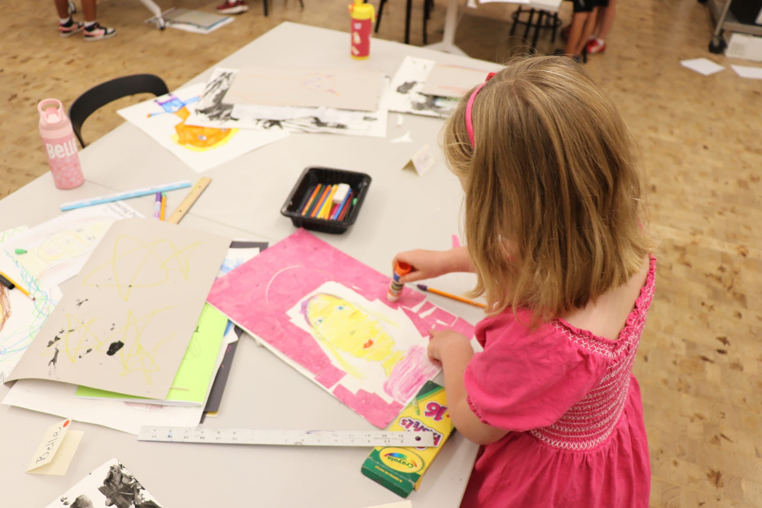 A young girl in a pink dress uses a glue stick while working on a colorful self-portrait at a table covered with art supplies, drawings, and crayons in a classroom or art studio.