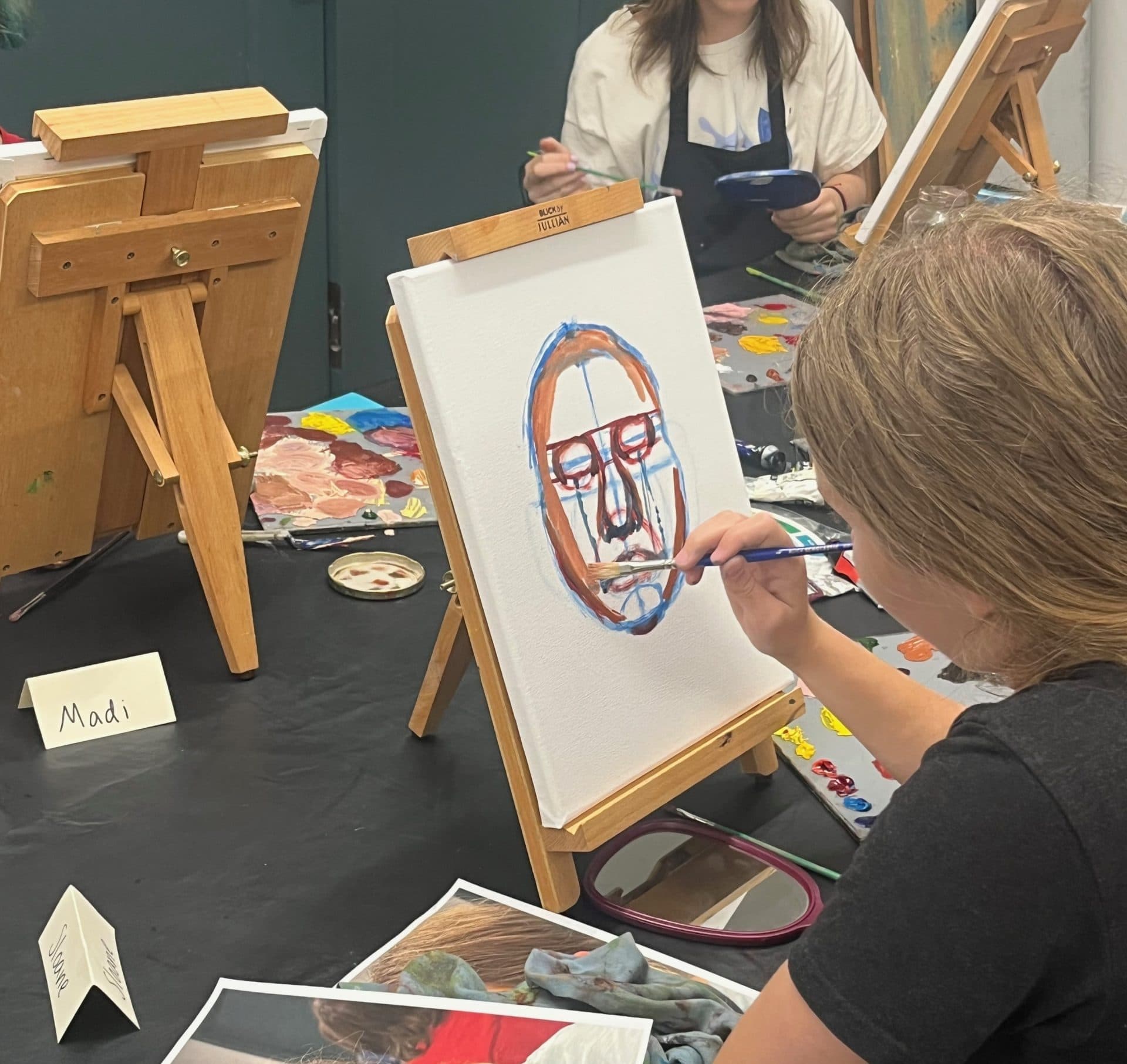 A person paints a colorful abstract face on a canvas at an art table, surrounded by paint supplies, reference photos, and easels. Another person works in the background. A name card labeled "Madi" is visible on the table.
