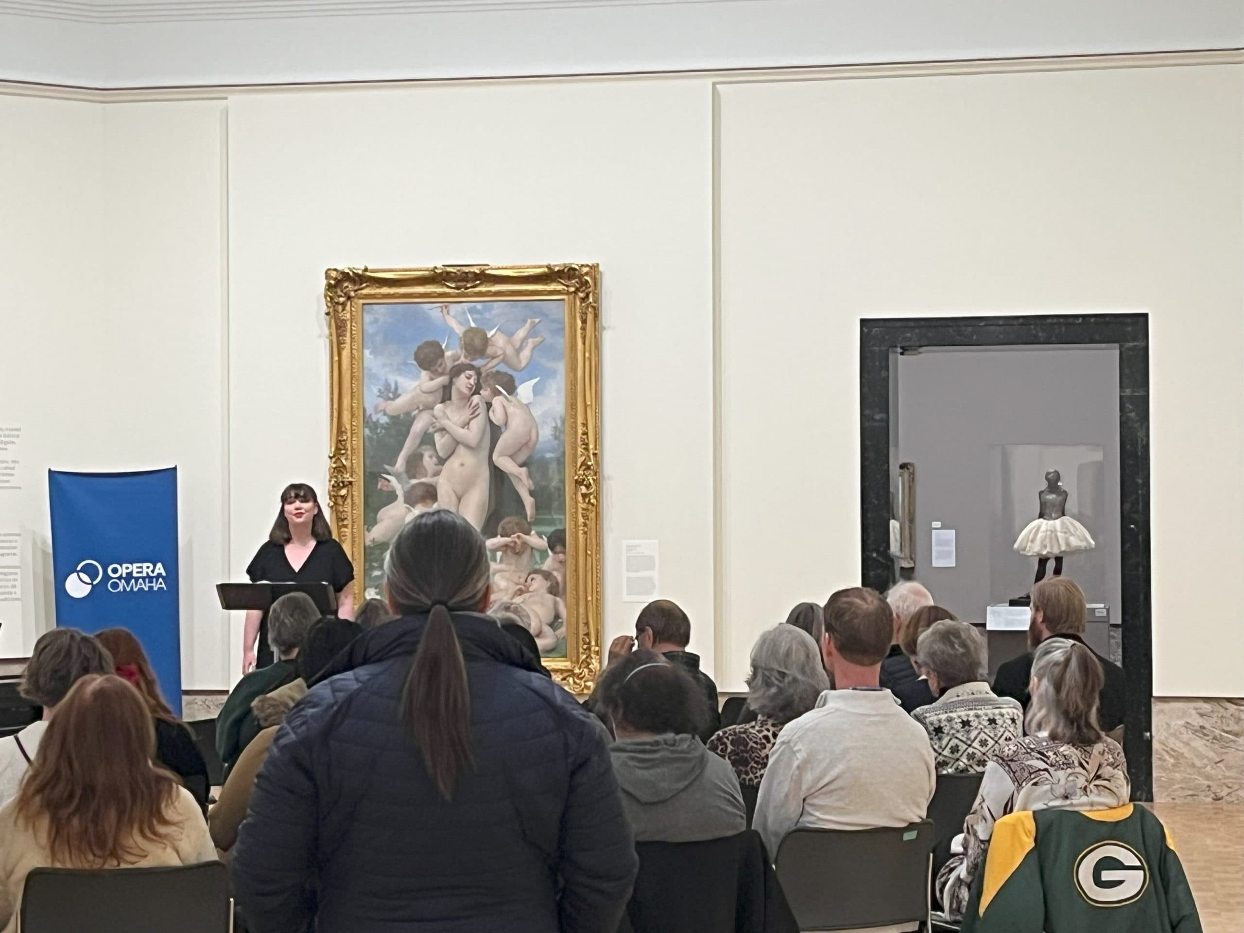 A woman stands in front of a seated audience, speaking in an art gallery. Behind her is a large classical painting, and to the side is a blue "Opera Omaha" banner. A sculpture is visible through a doorway.