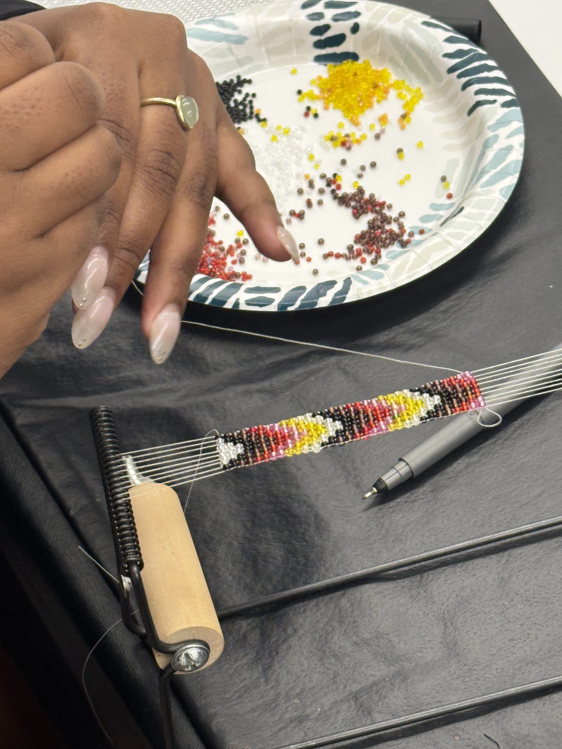 A person uses a bead loom to create a colorful bead pattern with yellow, red, white, and black beads. A paper plate with assorted beads sits nearby on a table.