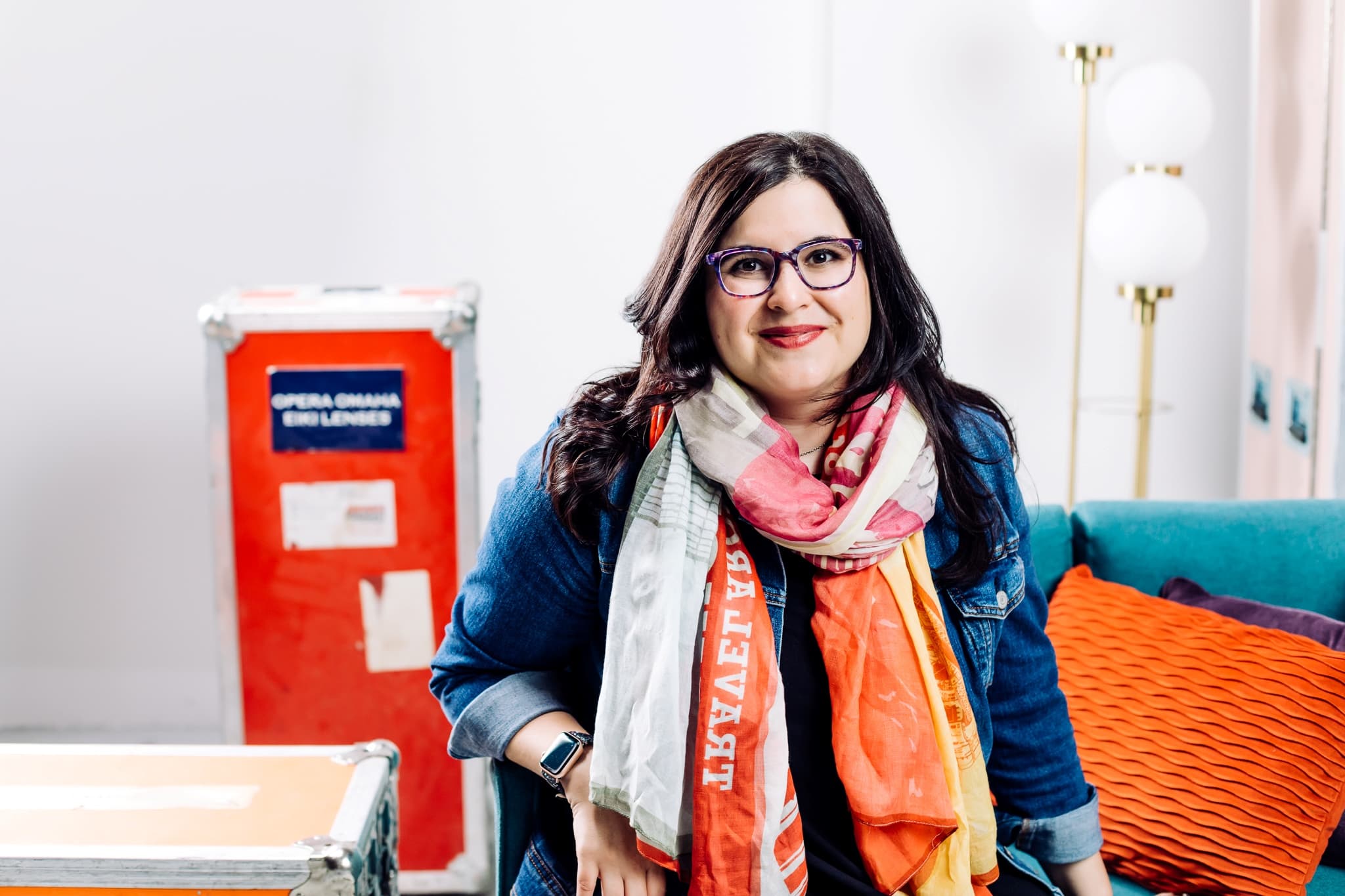 A woman with long dark hair and glasses sits on a teal couch. She wears a colorful scarf, denim jacket, and smiles at the camera. Behind her are an orange pillow, a red equipment case, and a floor lamp.