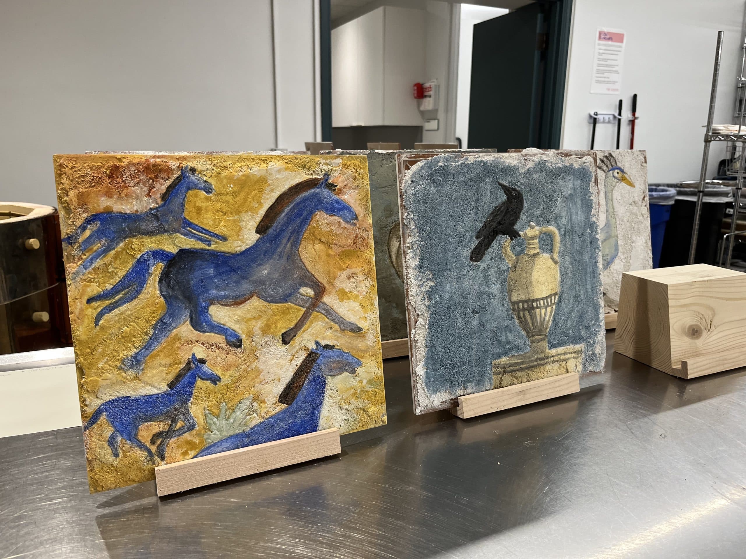Two painted tiles on wooden stands displayed on a metal surface. The left tile shows blue horses running, while the right tile depicts a black bird on a classical vase with another bird and partial object in the background.