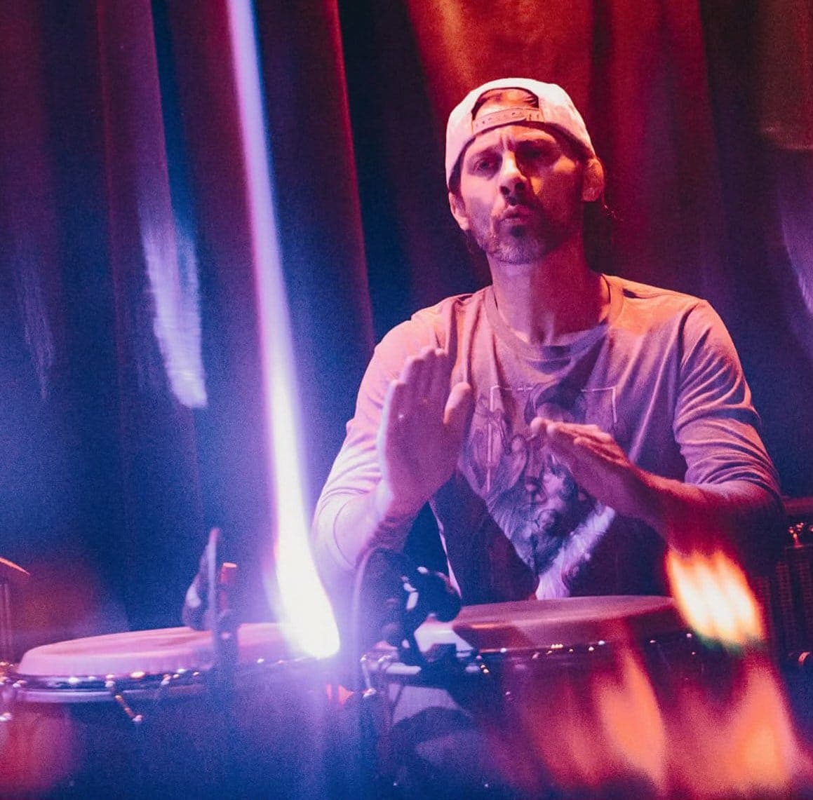 A man wearing a backward cap and casual shirt plays hand drums on stage under colorful, dramatic stage lighting, creating a vibrant and energetic atmosphere.