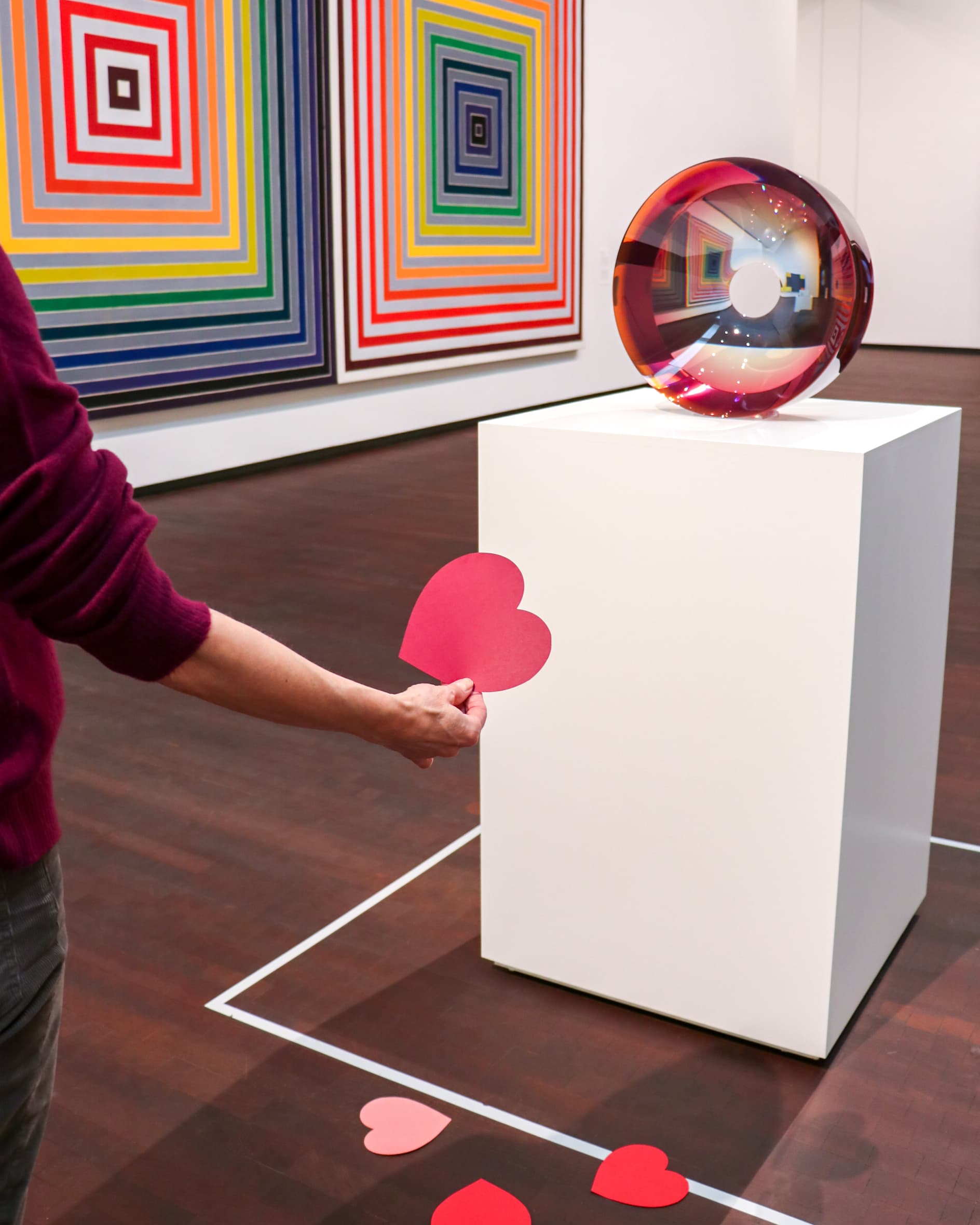A person holding a red paper heart stands near a white pedestal displaying a round, reflective sculpture in a gallery with colorful, geometric wall art. Red paper hearts are scattered on the floor.