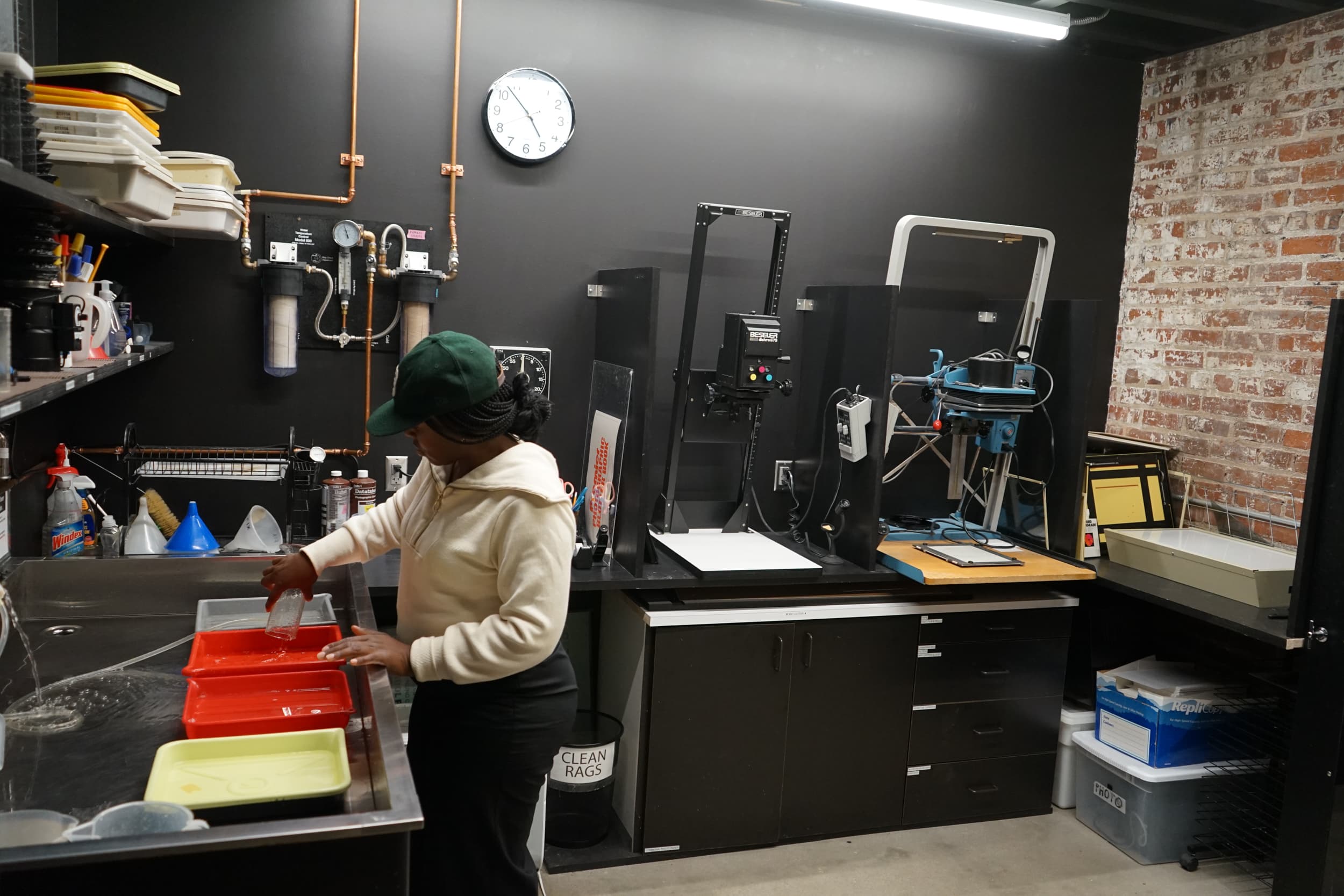A person in a white hoodie and green cap works in a darkroom, handling trays on a counter. The room has photo development equipment, a clock on the wall, shelves with supplies, and exposed brick.