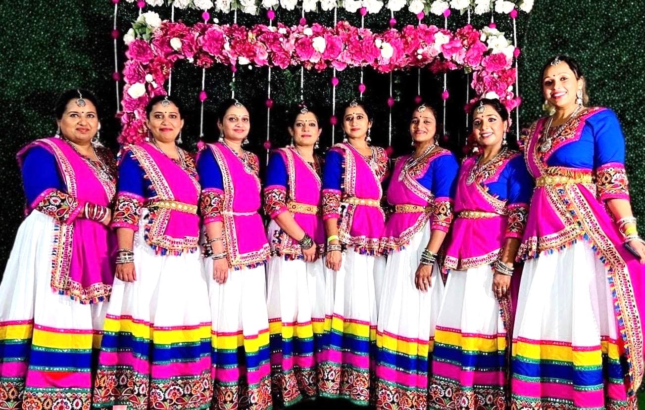 Eight women in traditional Indian attire, featuring blue and pink blouses with white skirts decorated with colorful borders, stand in a row, smiling, in front of a floral backdrop.
