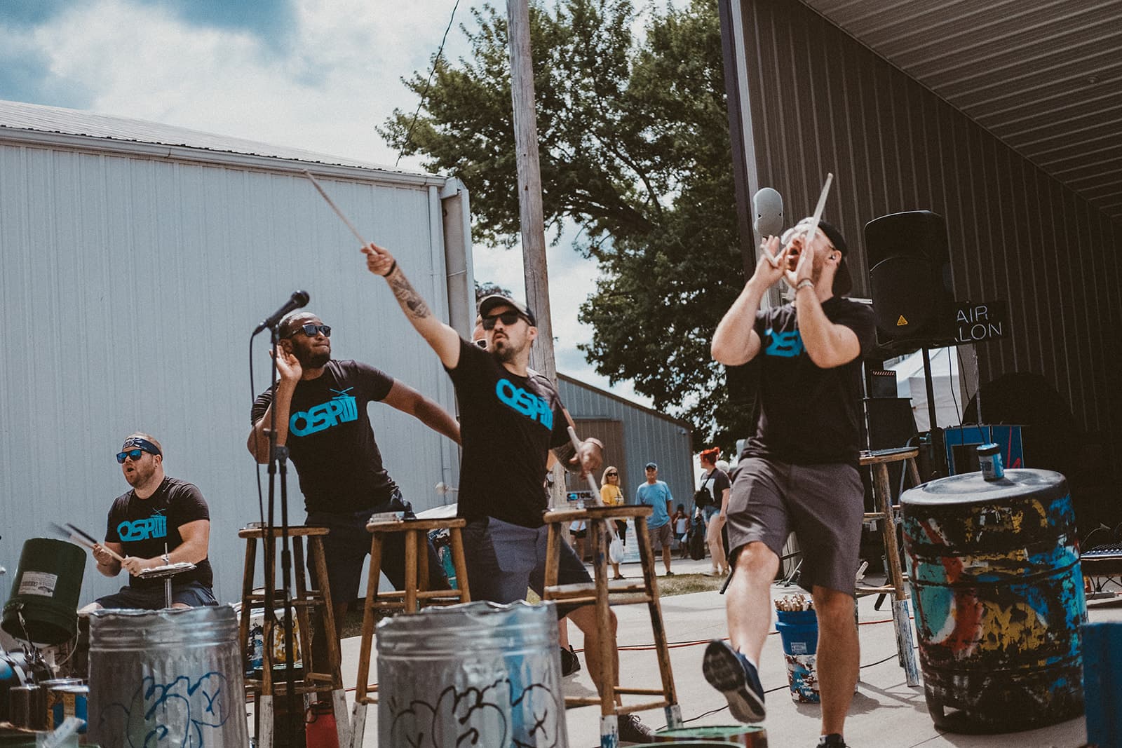 Three men performing an energetic drum routine outdoors, wearing matching black shirts and sunglasses, using metal trash cans as drums, with animated gestures. A small crowd and a large building are visible in the background.