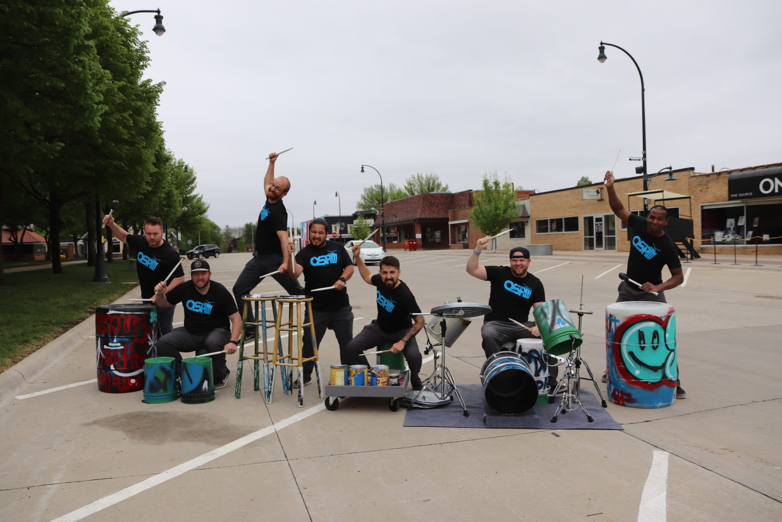 Six people in matching black Oseh shirts pose enthusiastically with drumsticks, ready to play drums made from buckets, barrels, and other recycled items on a deserted street, with shops and trees in the background.