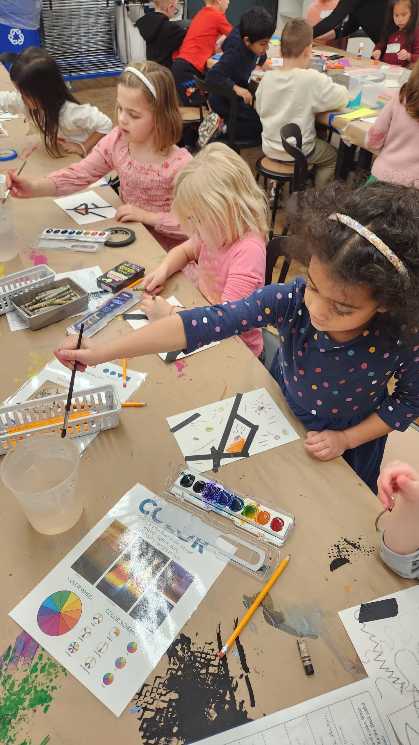 Young children sit around a table in a classroom, painting with watercolors. Art supplies and color charts are on the table, and the children appear focused on their creative projects.