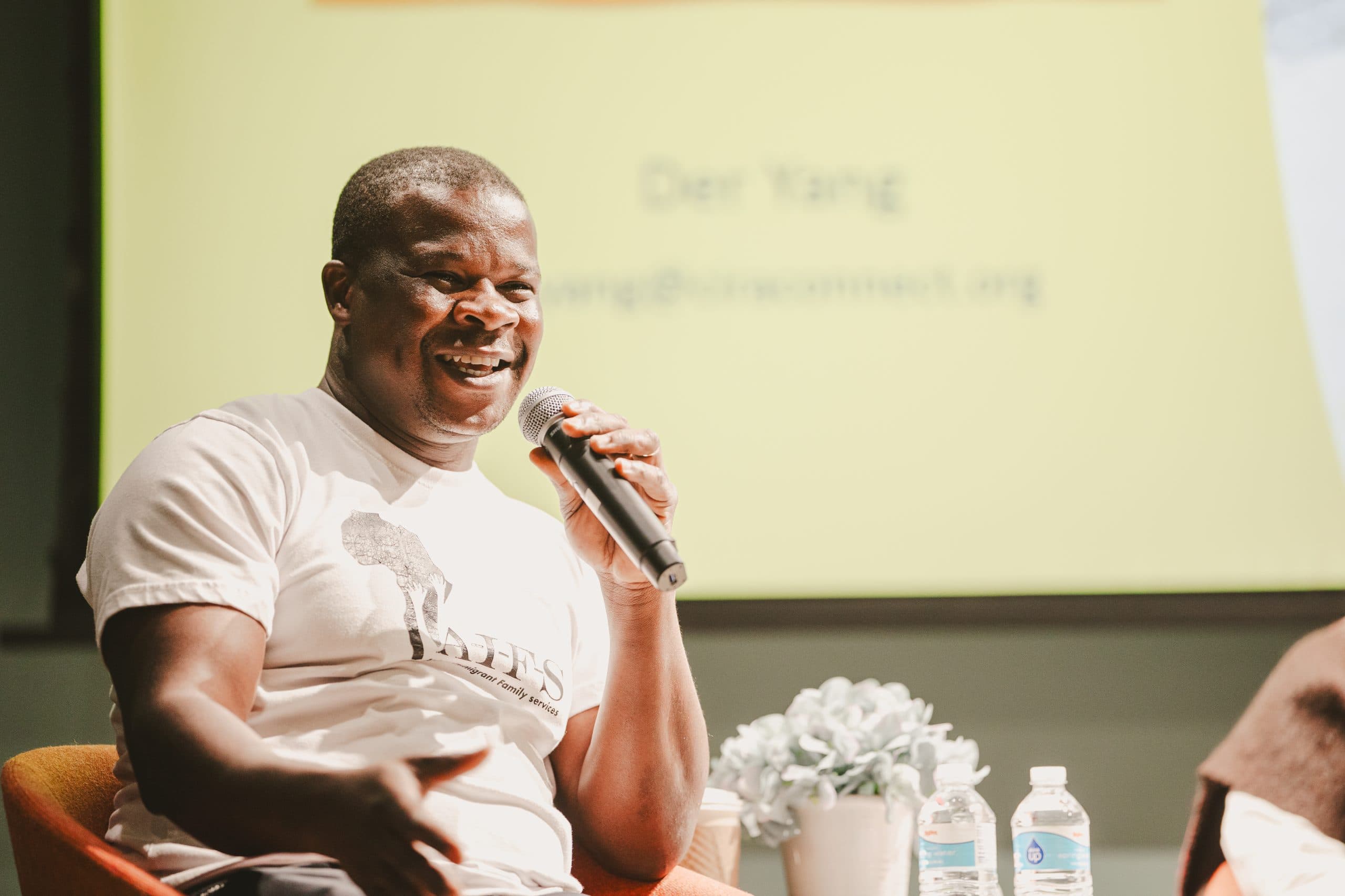 A man in a white T-shirt sits on an orange chair, speaking into a microphone. A yellow screen with text and two water bottles are visible in the background. A small plant sits on the table in front of him.