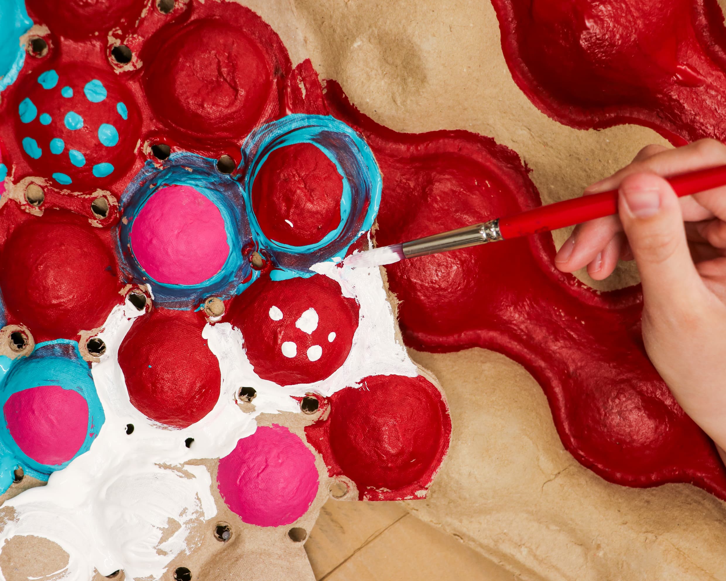 A hand uses a paintbrush to paint bright colors like red, blue, pink, and white on a textured cardboard egg carton, creating a colorful and artistic pattern.