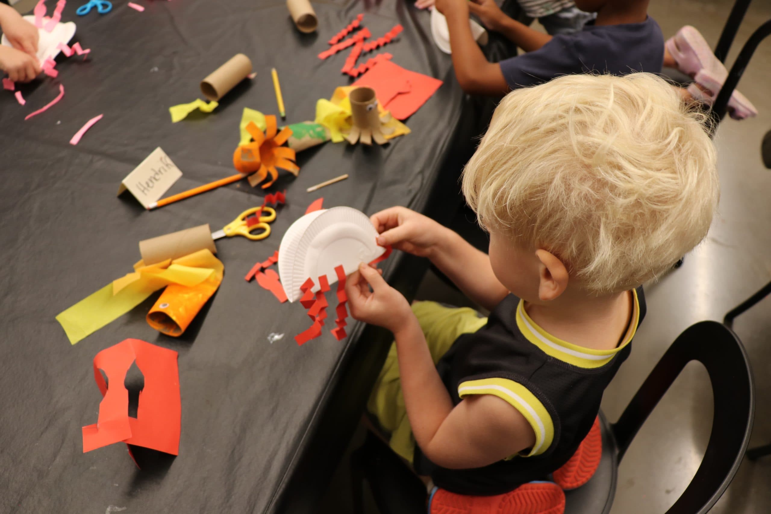 A young child with blond hair sits at a table making a craft with a paper plate and red paper strips. Various colorful craft supplies and tools, like scissors and cardboard tubes, are spread out on the table.