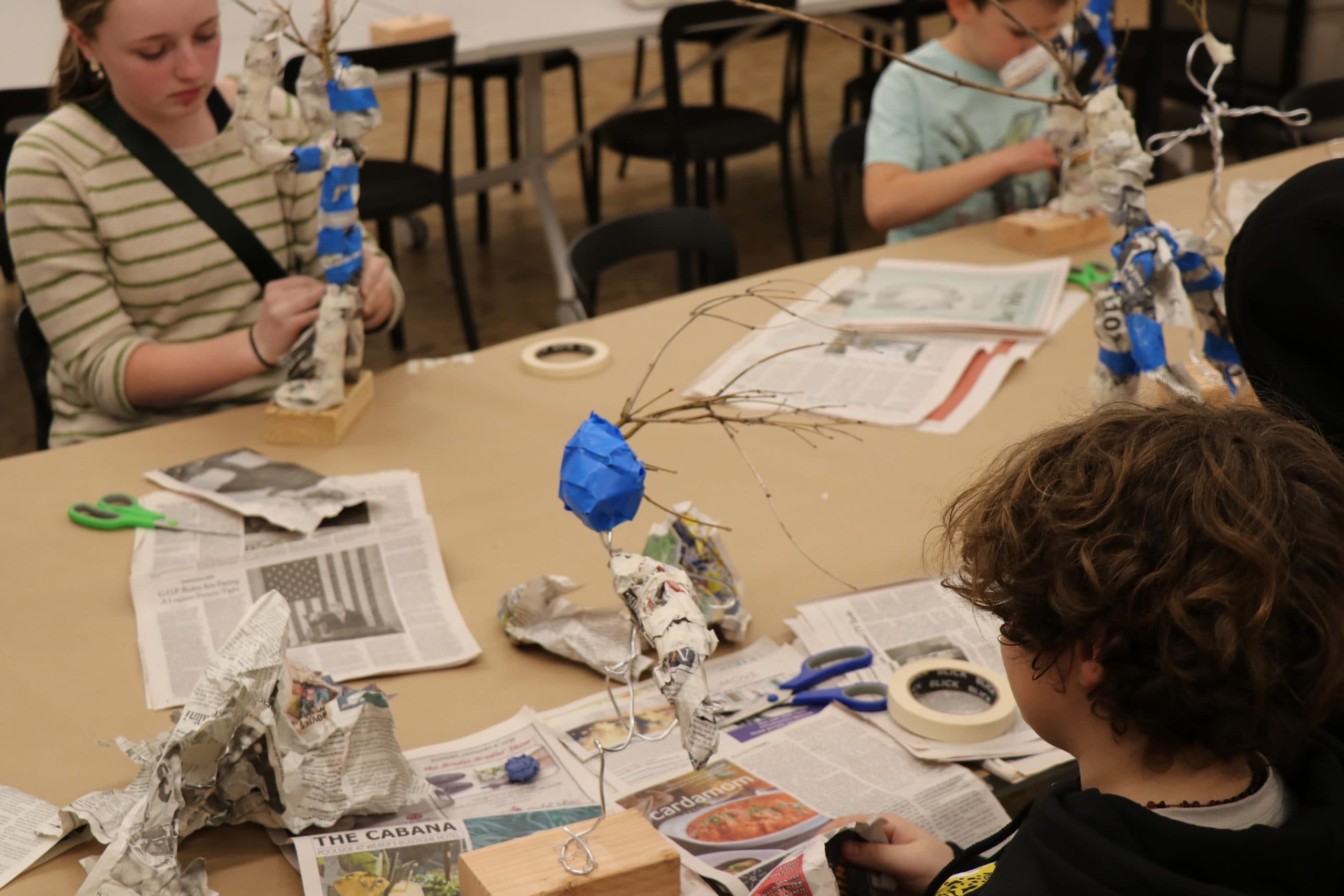 Children sit around a table covered in newspapers and art supplies, constructing sculptures with wire, tape, and paper. They concentrate on building tree-like structures mounted on wooden bases.