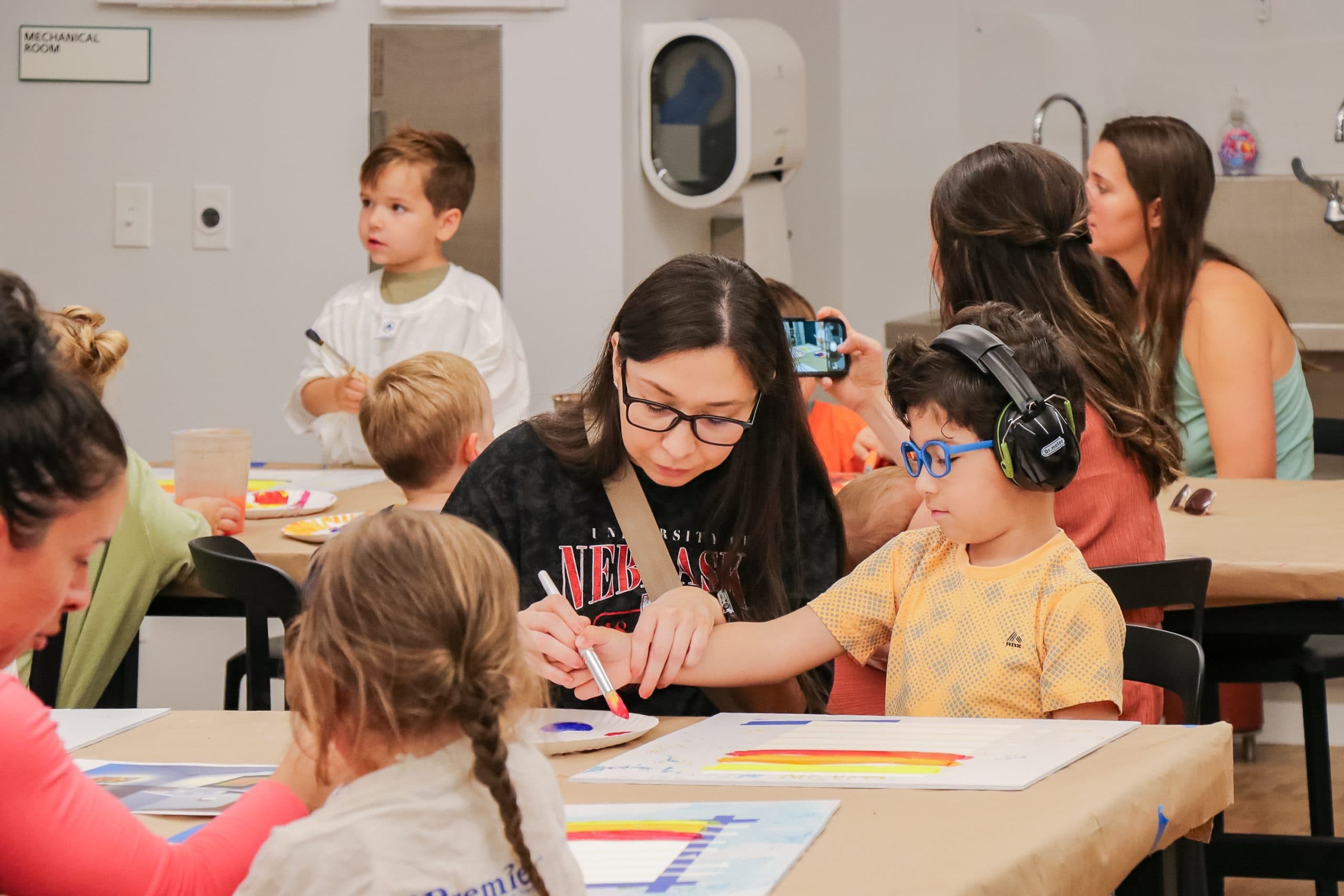 Children and adults sit at tables painting on paper. One child wears headphones and blue glasses, while an adult helps him with his artwork. Other children and adults engage in similar activities in the background.