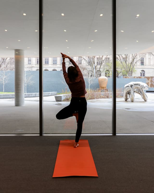 A person practices yoga on an orange mat indoors, standing in tree pose facing large floor-to-ceiling windows with a cityscape and outdoor sculptures visible outside.