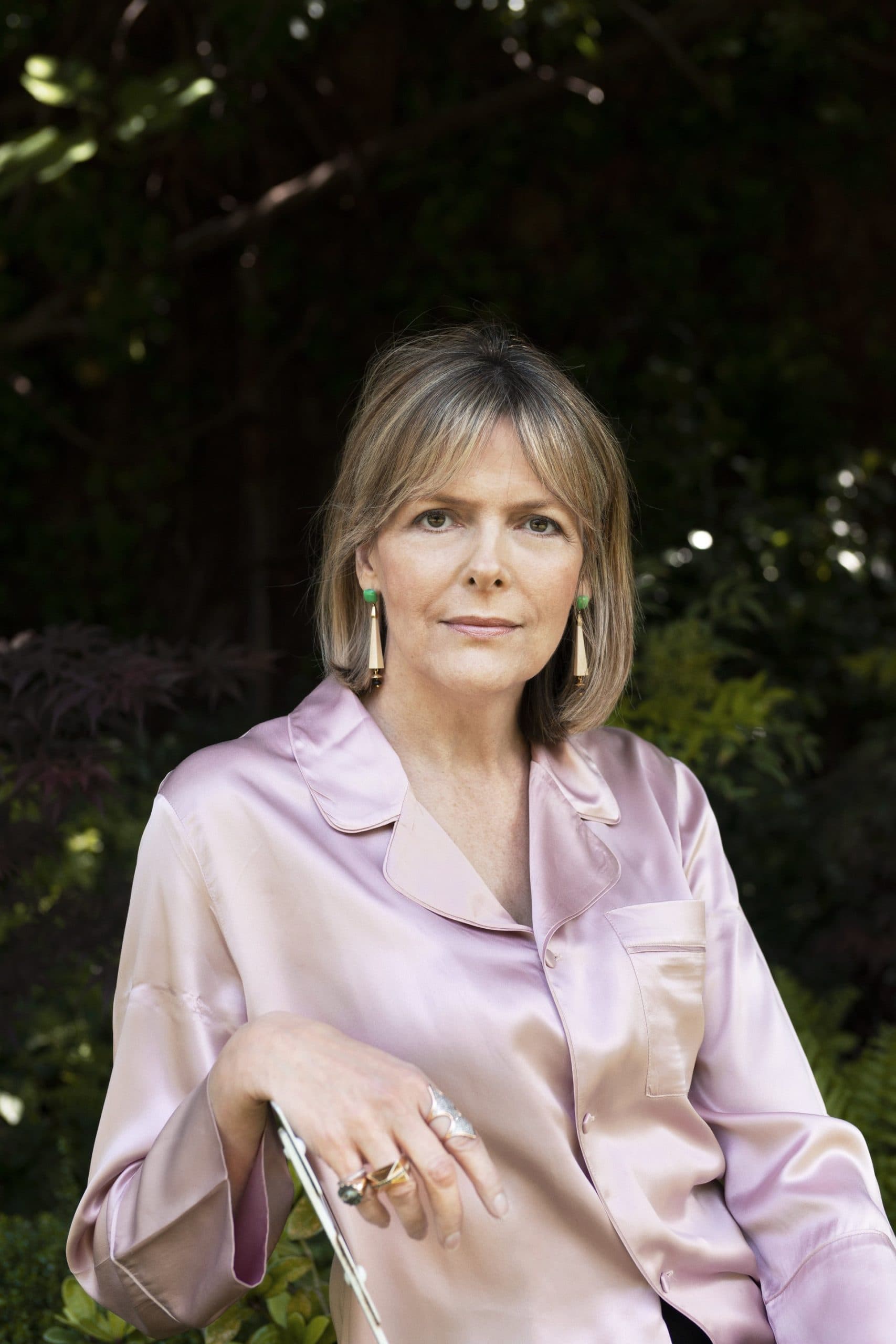 A middle-aged woman wearing a pale pink button-up shirt and dangling earrings sits outdoors.