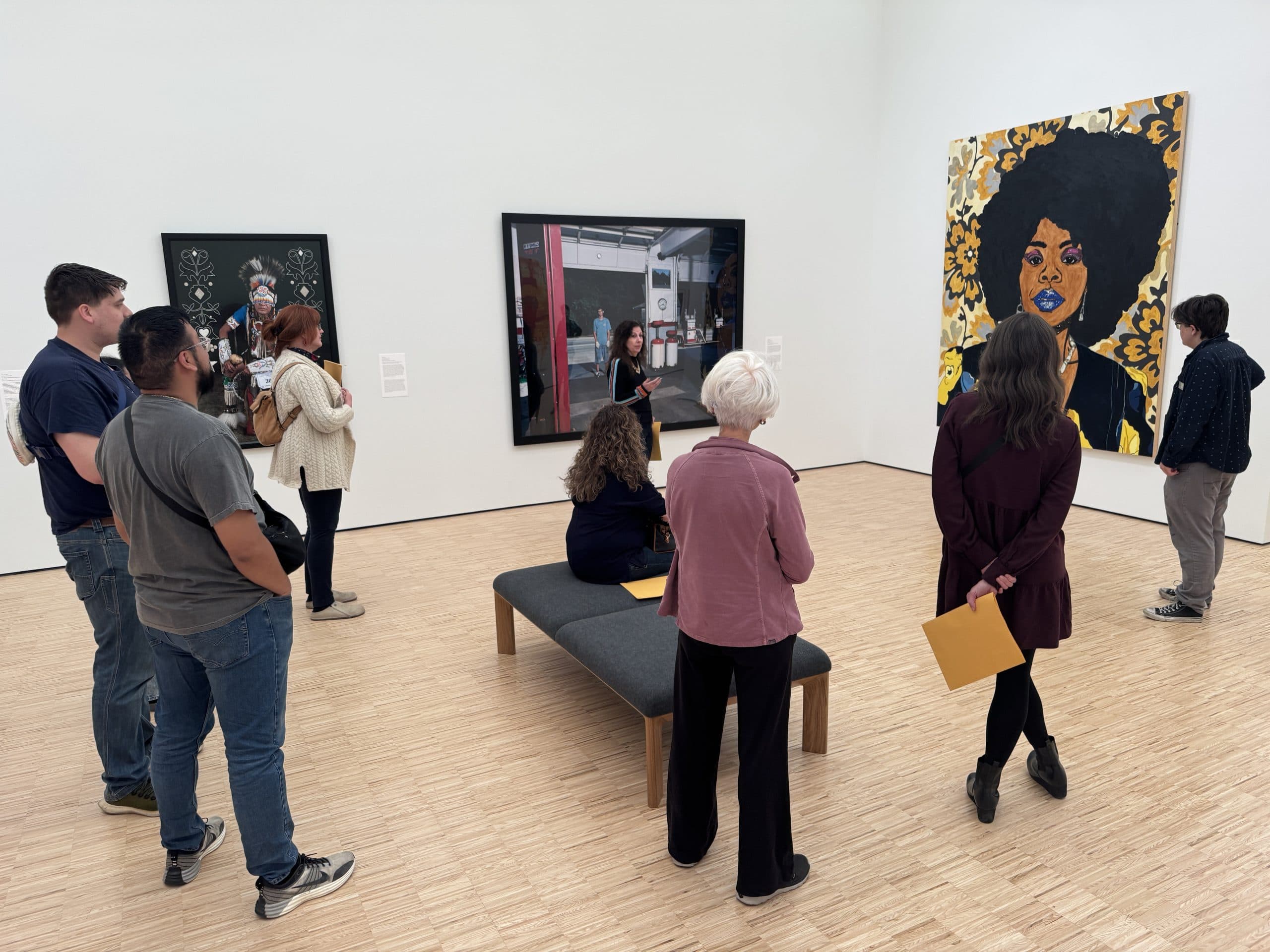 A group of people observes various artworks in a bright, modern gallery. One person sits on a bench while others stand. Art includes a large portrait of a woman with an afro on the right wall.
