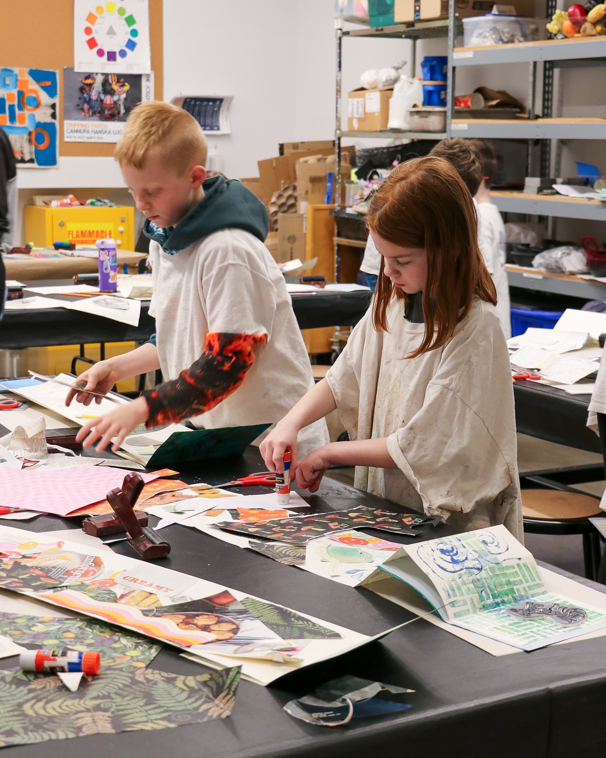 Two children in an art classroom use glue sticks to create colorful collages with paper cutouts on a table covered in art supplies. Shelves with bins and art projects are visible in the background.