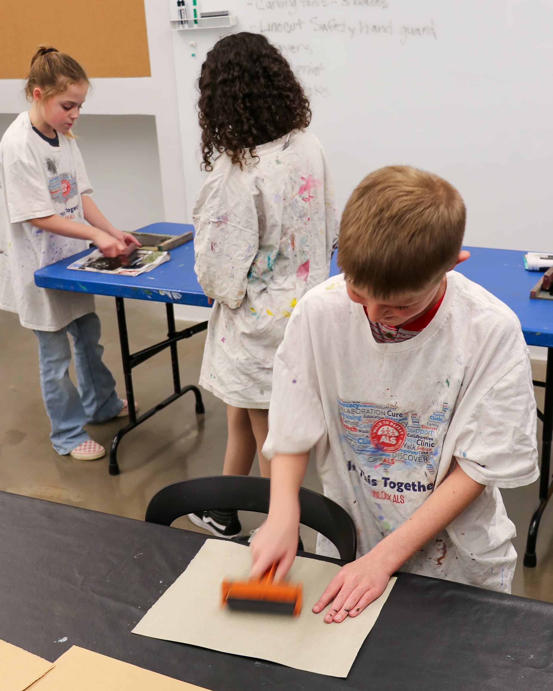 Three children in paint-splattered shirts use brayers to roll ink onto paper during an art class.