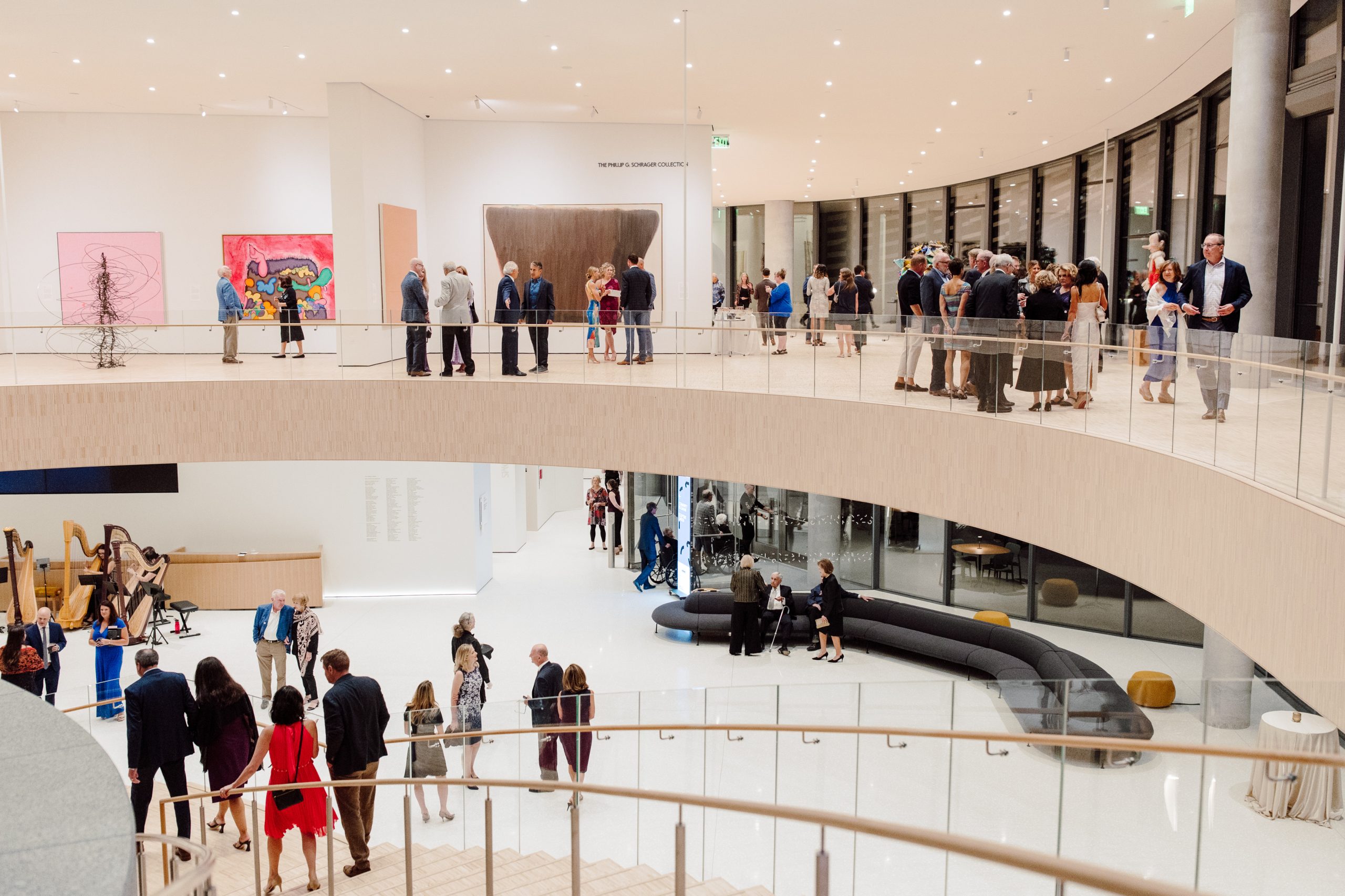 People gather and converse on two levels of a modern art gallery. The upper level features colorful artworks on the walls, while the lower level includes seating and a small group near a harp player. Bright lighting and clean design are prominent.