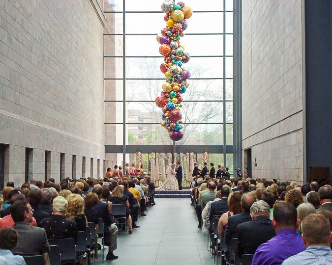 A wedding ceremony takes place in a large hall with a tall glass window. Guests are seated on both sides, facing the couple and officiant. Colorful balloons are suspended from the ceiling, adding a festive touch to the neutral-toned space.