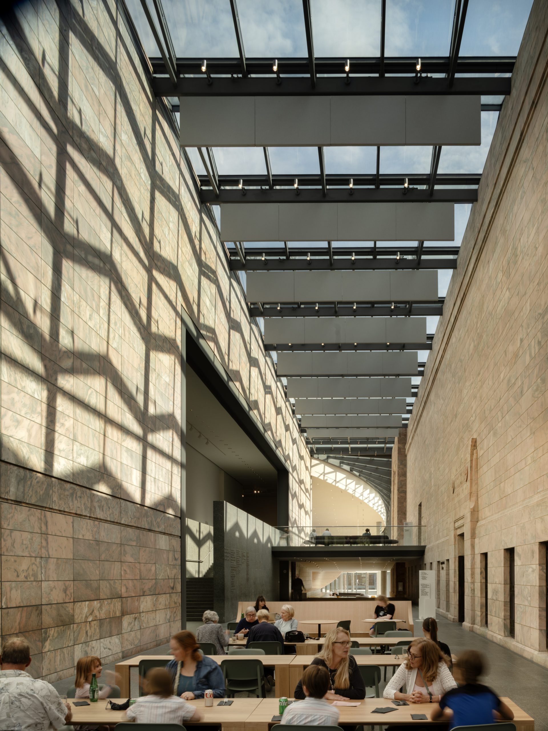 People sitting at tables inside a large, modern atrium with high glass ceilings and stone walls. The space is well-lit with natural light, and geometric shadows are cast on one wall.